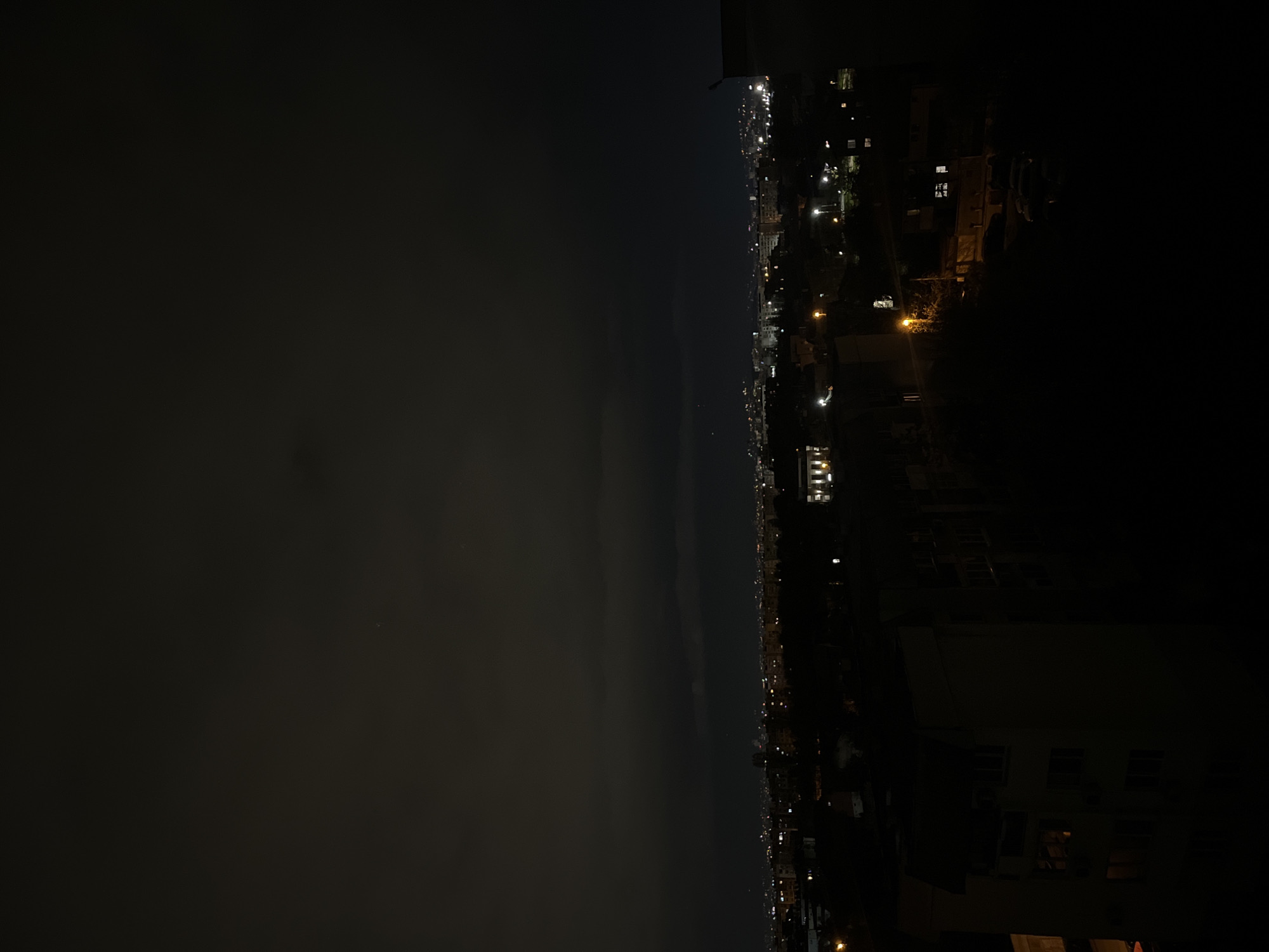 The image is a nighttime cityscape, possibly in Sofia, Bulgaria, based on the architecture and street layout. The left side of the image is dominated by a dark, cloudy sky. On the right, illuminated buildings, houses, and streets with sporadic lights outline a portion of the city. The lights vary in intensity, creating a visual depth. The image is taken from a high vantage point, possibly a building or a hill overlooking the city. The overall scene suggests a quiet night with urban lighting juxtaposed against the natural darkness.