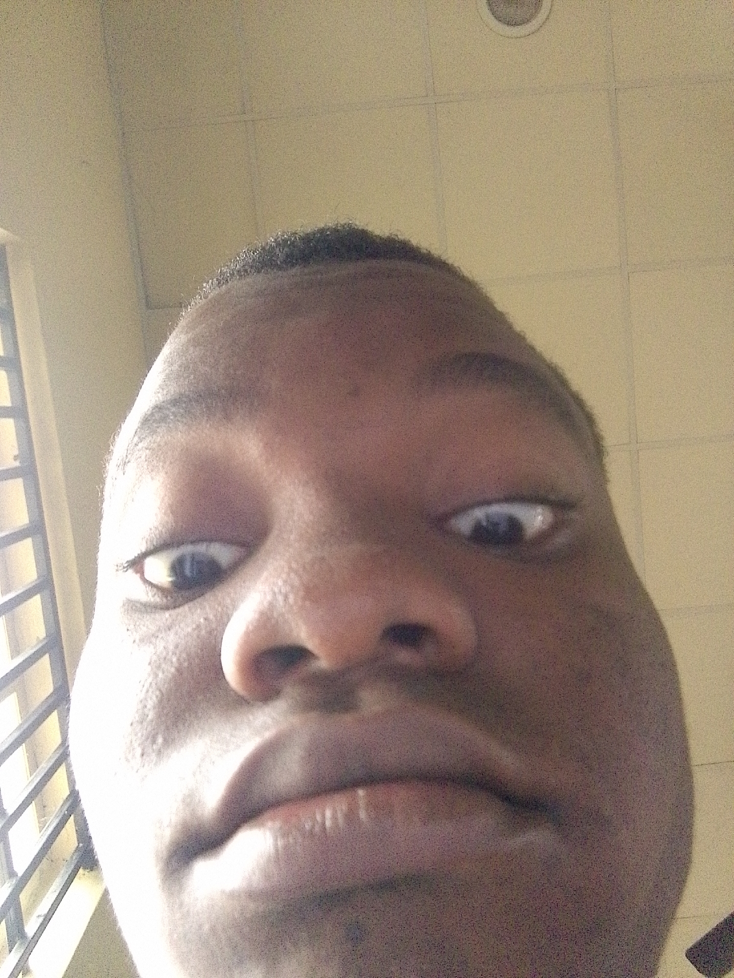 A close-up, low-angle selfie captures the face of a young person of African descent, filling the frame. The subject's eyes are wide and looking directly at the camera. Their nose is prominent, and their lips are slightly parted. The skin texture is visible, with some pores and a slight sheen. The background appears to be an indoor ceiling with tiled panels and a circular light fixture. To the left of the subject, a barred window with vertical and horizontal elements is partially visible, suggesting an interior space with an external view. The lighting is bright, casting some shadows. The image is grainy, indicating it was taken with a low-resolution camera or in less than ideal lighting conditions. The subject's head is tilted, and the perspective is distorted due to the close proximity of the camera and the wide-angle lens effect. The location is identified as Eleweran, Nigeria, suggesting an urban or semi-urban environment.