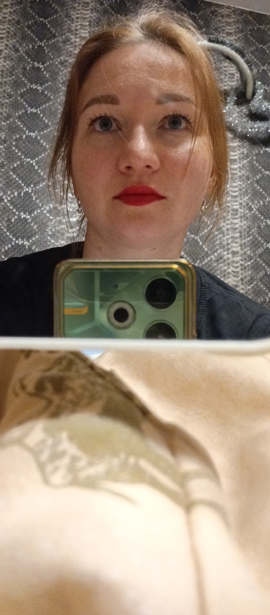 The image is a selfie of a woman with fair skin, reddish-blonde hair, and light blue eyes. She has on red lipstick and is wearing earrings. She is holding up a cell phone, with its camera and a reflection of a light-colored surface visible in the foreground. The background is a textured gray pattern. The location appears to be indoors, but the specific city or area is unknown.