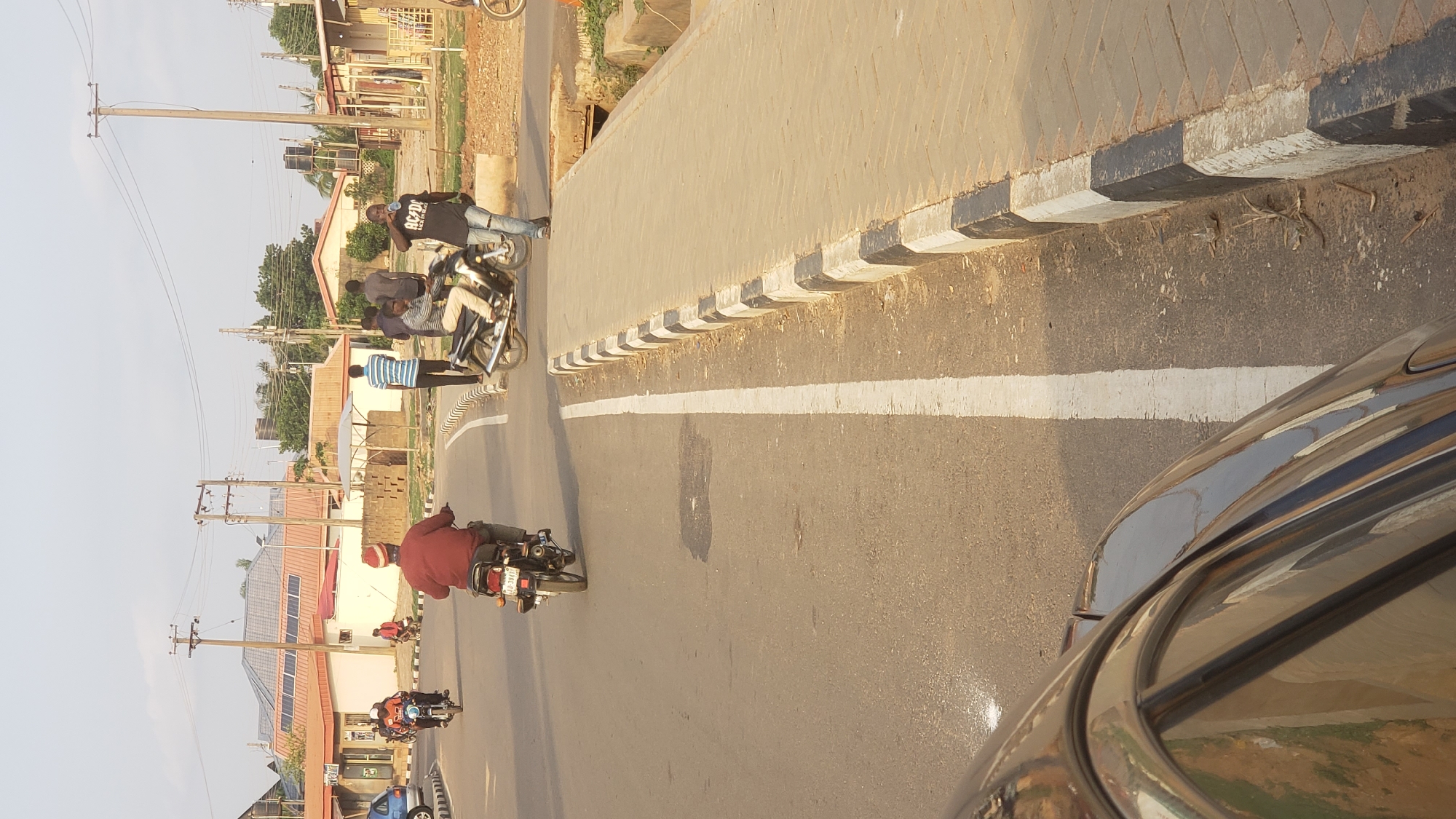 The image depicts a street scene in Narayi, Nigeria. The road is paved with a white dividing line. A motorcycle is driving away from the viewer. A group of men are standing by the sidewalk; one man is on a motorcycle. Houses are visible in the background. Power lines and utility poles line the side of the road. A car is visible in the right corner of the image.