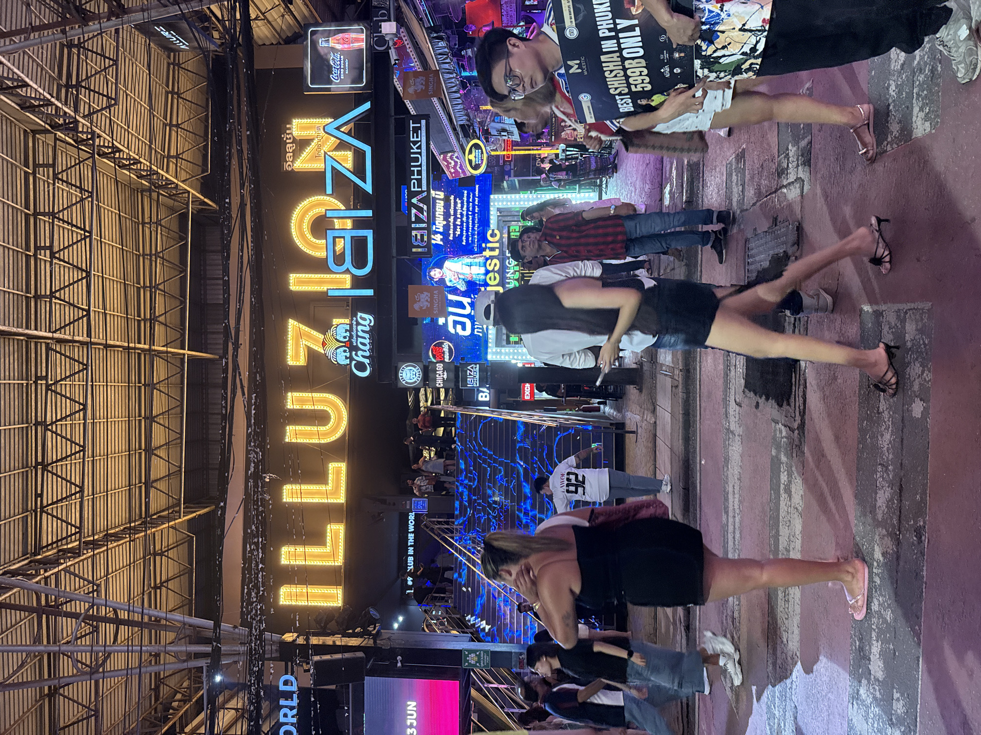 The image depicts a bustling street scene at night, illuminated by vibrant neon signs and overhead lighting. Dominating the scene is a large, illuminated sign that reads "ILLUZION IBIZA," with "ILLUZION" in bright yellow and "IBIZA" in blue. Below this, smaller signs indicate "IBIZA PHUKET" and a Coca-Cola advertisement.
A group of people are walking across a pedestrian crossing in the foreground. To the left, a woman in a black dress and pink flip-flops walks away from the camera. Beside her, a woman in a white top and denim shorts is walking. Further to the left, a man in a blue shirt and jeans is walking.
In the background, a set of blue-lit stairs leads up to a club entrance. People can be seen standing on the stairs and at the top. Several other individuals are walking around the area, some looking towards the camera, others engaged in their own activities. The environment appears to be a lively entertainment district, likely a tourist area, given the signage and the diverse crowd. The overall impression is one of a vibrant nightlife atmosphere.