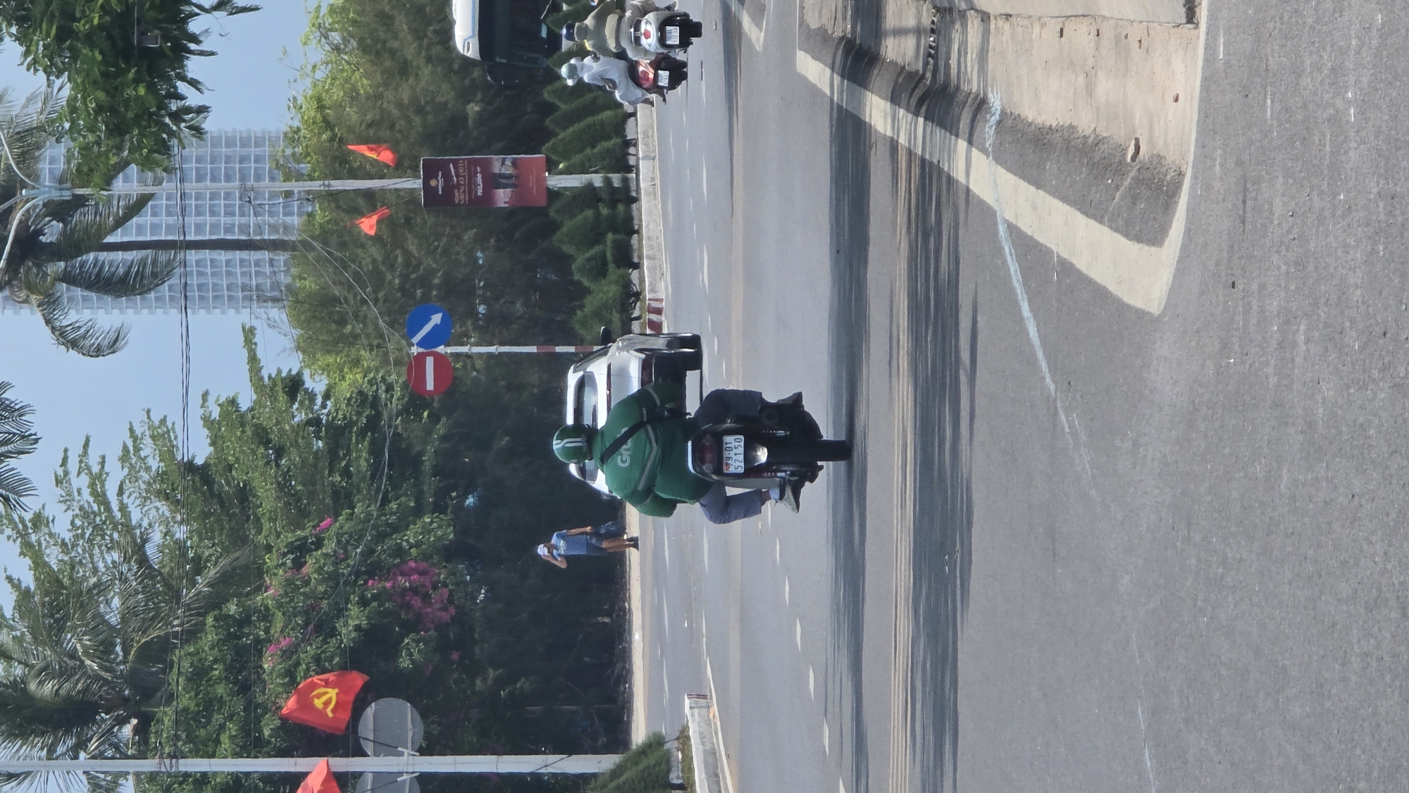 The image depicts a street scene in Nha Trang, Vietnam. A person wearing a green Grab uniform is riding a motorcycle with license plate "79-D1 521.50" on a road. There are other motorcycles and vehicles in the background. The environment includes trees, a building in the distance, and traffic signs, including a no entry sign and a one way sign. There are also red flags.