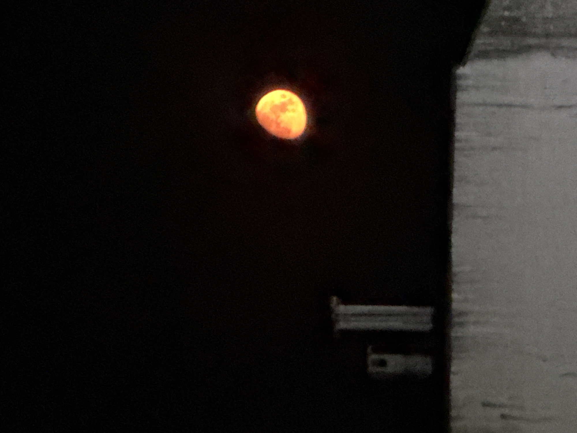 The image shows a full moon, orange in color, against a dark sky. To the right of the frame, there's a blurred, out-of-focus portion of a building or structure, possibly made of concrete or similar material. Below the building, there are faint shapes that resemble a pipe or fitting. The background is entirely dark, with no other discernible details. Based on the image content alone, it is not possible to determine whether the image was taken in Varna, Bulgaria.