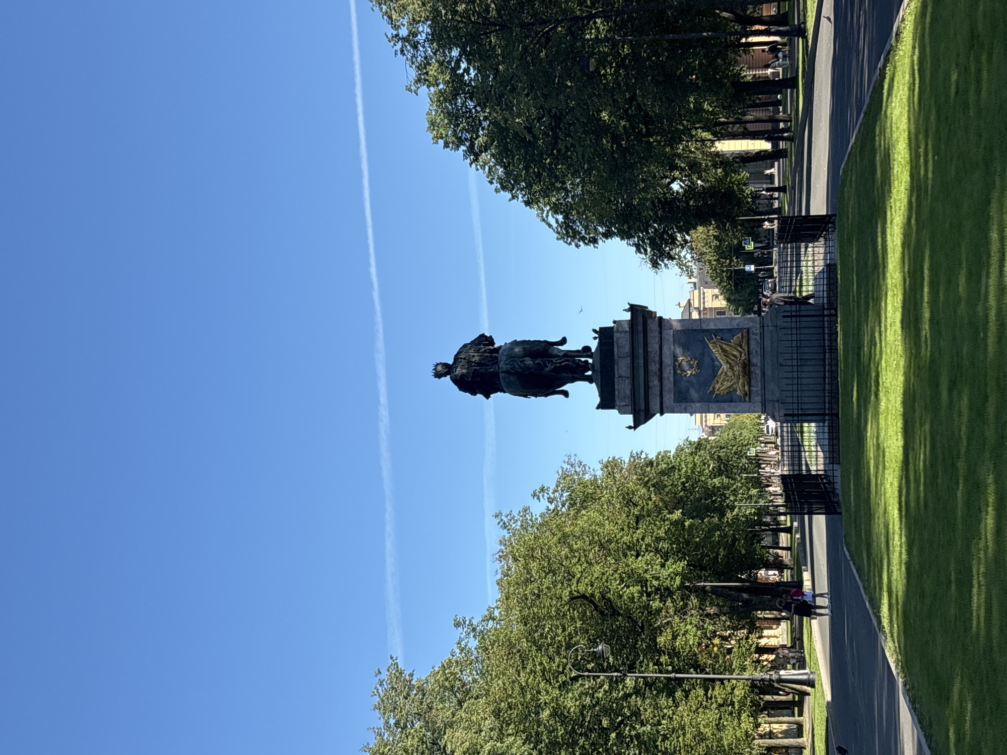 The image captures an equestrian statue in a park-like setting under a clear blue sky with faint contrails. The statue, made of dark metal, depicts a person wearing a crown, seated on a horse. The statue stands on a substantial, ornate pedestal with gold detailing. The park features a well-maintained lawn in the foreground and a paved pathway lined with mature trees on either side. A black metal fence encloses the area around the statue. Several people are visible walking on the pathway in the background, dressed casually. Streetlights are interspersed among the trees. In the distance, buildings with classical architecture are visible. The overall atmosphere is serene and historic, suggesting a public monument in a significant urban green space in Saint Petersburg, Russia, likely the Bronze Horseman, or a similar monument in the city.