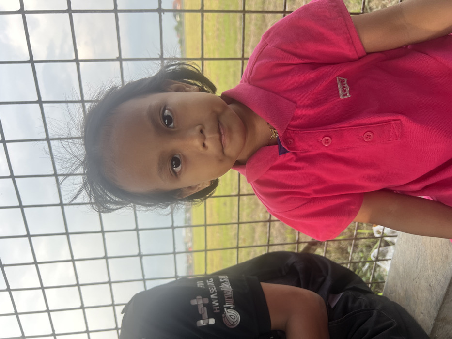 A young girl with dark hair and eyes is seen from the chest up, wearing a bright pink polo shirt with a Levi's logo on the left side. She stands behind a wire fence, partially obscuring a grassy field with scattered buildings in the background. A portion of a person wearing a black shirt with white writing is visible below the girl. The image has a slightly rotated orientation.