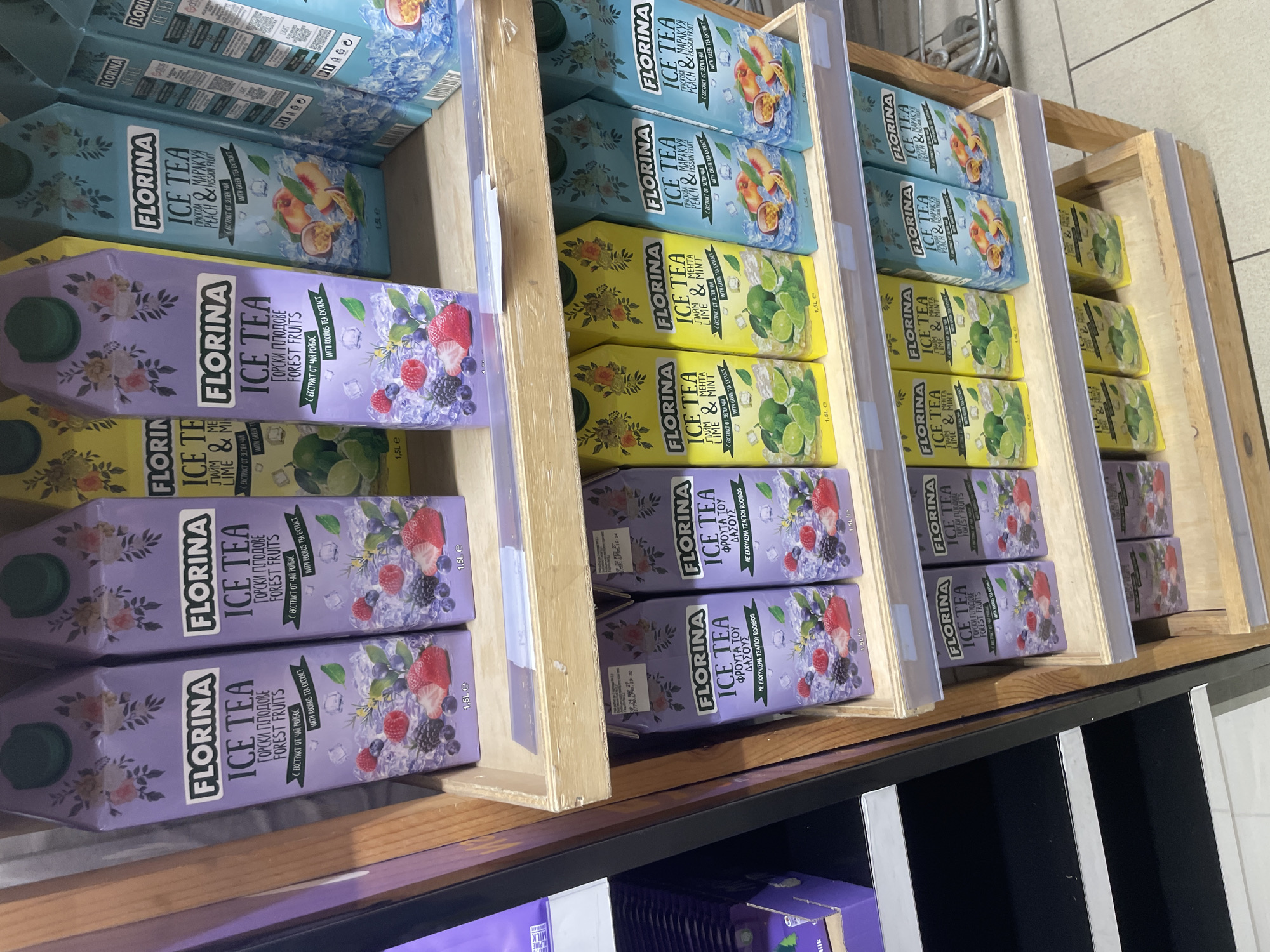 This image captures a retail display of Florina Ice Tea products in what appears to be a supermarket or grocery store in Ihtiman, Bulgaria. The ice tea cartons are neatly arranged in multi-tiered wooden crates, showcasing a variety of flavors.
The primary subjects are numerous 1.5-liter cartons of "FLORINA ICE TEA." Three distinct flavors are visible:
1. **Peach & Passion Fruit:** Featured on light blue cartons, with an illustration of peaches, passion fruit, and ice cubes. The packaging states "PEACH & PASSION FRUIT" (ПРАСКОВА & МАРАКУЯ) and "WITH GREEN TEA EXTRACT" (С ЕКСТРАКТ ОТ ЗЕЛЕН ЧАЙ).
2. **Lime & Mint:** Presented on yellow cartons, depicting limes, mint leaves, and ice cubes. The text reads "LIME & MINT" (ЛАЙМ & МЕНТА) and "WITH GREEN TEA EXTRACT" (С ЕКСТРАКТ ОТ ЗЕЛЕН ЧАЙ).
3. **Forest Fruits:** Shown on purple cartons, illustrating a mix of berries like strawberries, raspberries, blueberries, and blackberries with ice. The packaging identifies the flavor as "FOREST FRUITS" (ГОРСКИ ПЛОДОВЕ) and indicates "WITH ROOIBOS TEA EXTRACT" (С ЕКСТРАКТ ОТ ЧАЙ РОЙБОС). Some cartons also display the Greek translation: "ΦΡΟΥΤΑ ΤΟΥ ΔΑΣΟΥΣ" and "ΜΕ ΕΚΧΥΛΙΣΜΑ ΤΣΑΓΙΟΥ ROOIBOS."
The display is well-stocked, with multiple rows and columns of each flavor. The cartons are standard brick-shaped containers with green screw caps. The background shows parts of other store shelving and a tiled floor, suggesting an indoor retail environment. A label reading "Milk" is partially visible on a product at the bottom left, indicating other items are sold nearby. The Bulgarian text on the packaging aligns with the provided location context of Ihtiman, Bulgaria.