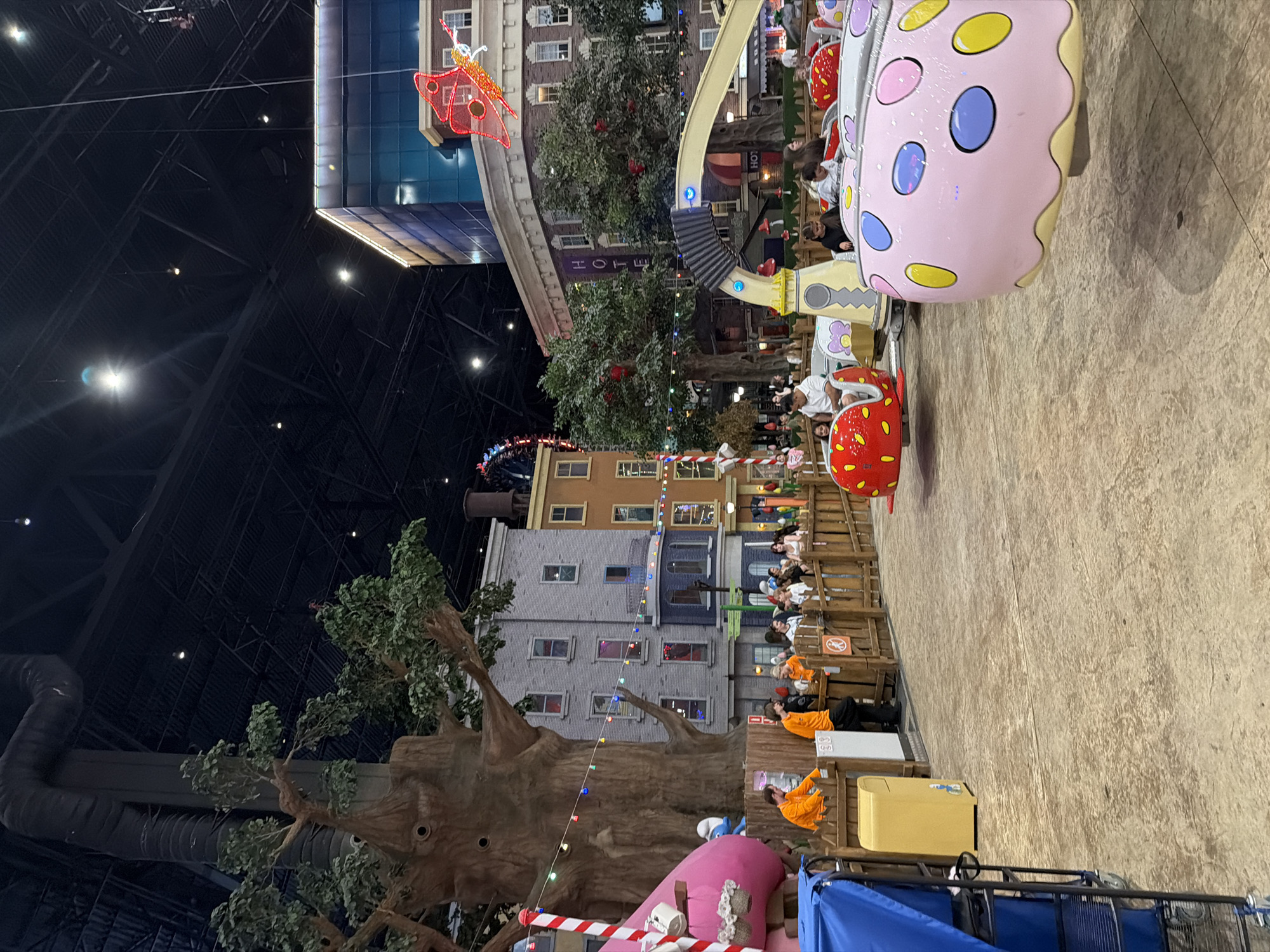 An indoor amusement park attraction features a whimsical, cartoon-like environment. In the foreground, several small, brightly colored ride vehicles, shaped like strawberries and donuts, are in motion. One pink donut-shaped vehicle with colorful dots is prominently displayed. A yellow trash can is visible near a wooden fence where several park employees in orange shirts are standing. In the background, faux buildings resembling a city street are set up, complete with a large, artificial tree. Overhead, the dark expanse of the indoor venue's ceiling is visible, with exposed beams and lighting fixtures. Several people, including families and children, are enjoying the rides. The overall atmosphere suggests a lively and entertaining space designed for children.