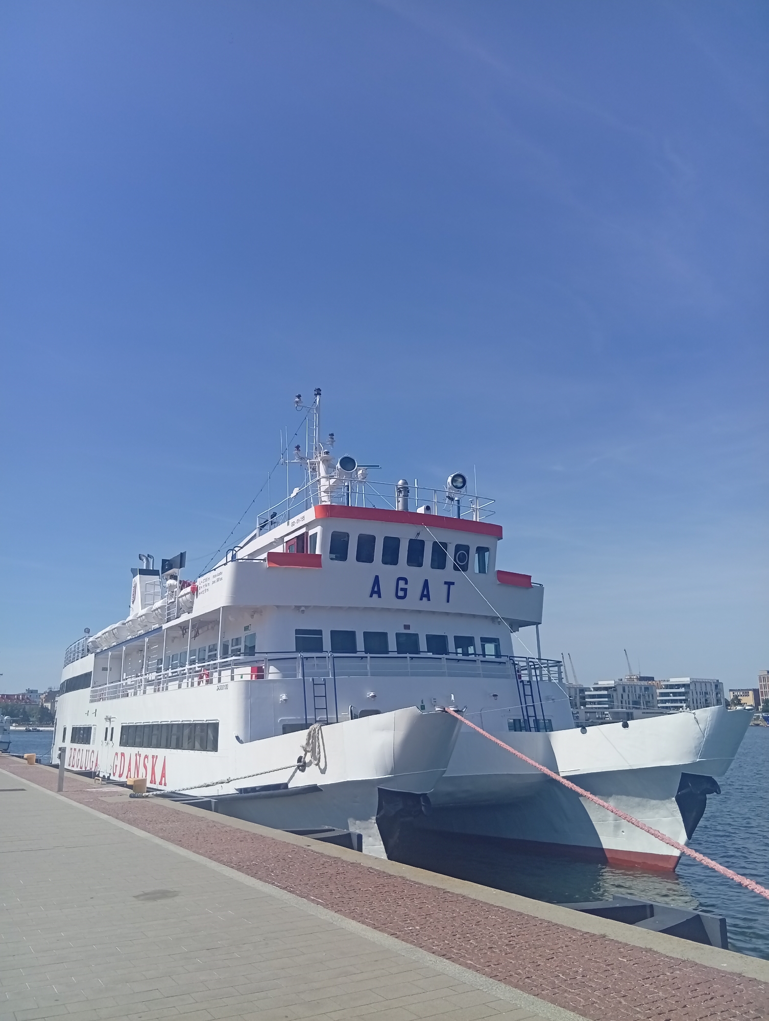 A white catamaran ferry named "AGAT" is docked at a pier in Gdynia, Poland. The ferry has red trim along its upper decks and the name "ZEGLUGA GDANSKA" written in red lettering on its side. The vessel is secured with thick ropes to the pier, which is paved with light gray and reddish-brown interlocking stones. The sky is clear blue with a few wispy clouds. In the background, modern buildings and cranes are visible, suggesting an urban waterfront area. The water is calm and reflects the blue of the sky. There are no people visible in the image.