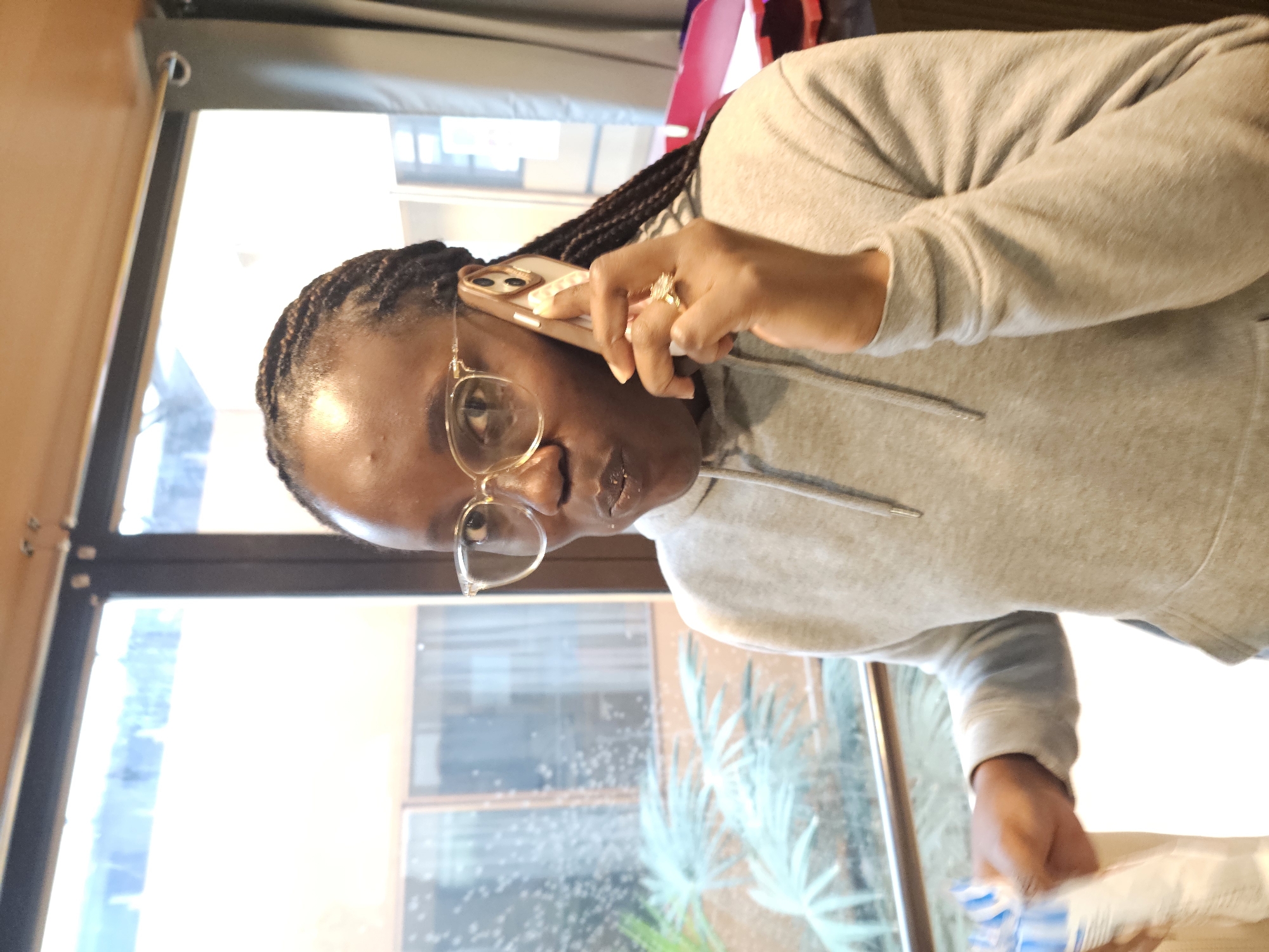 A woman with brown braided hair, wearing wire-rimmed glasses and a gray hooded sweatshirt, is on a phone call. She is holding the phone to her left ear with her left hand, which has a prominent ring on the ring finger. The background is blurred, showing a window with outdoor greenery and a glimpse of a red or pink object. The location is not definitively identifiable as Paris, France, from the visual information provided.