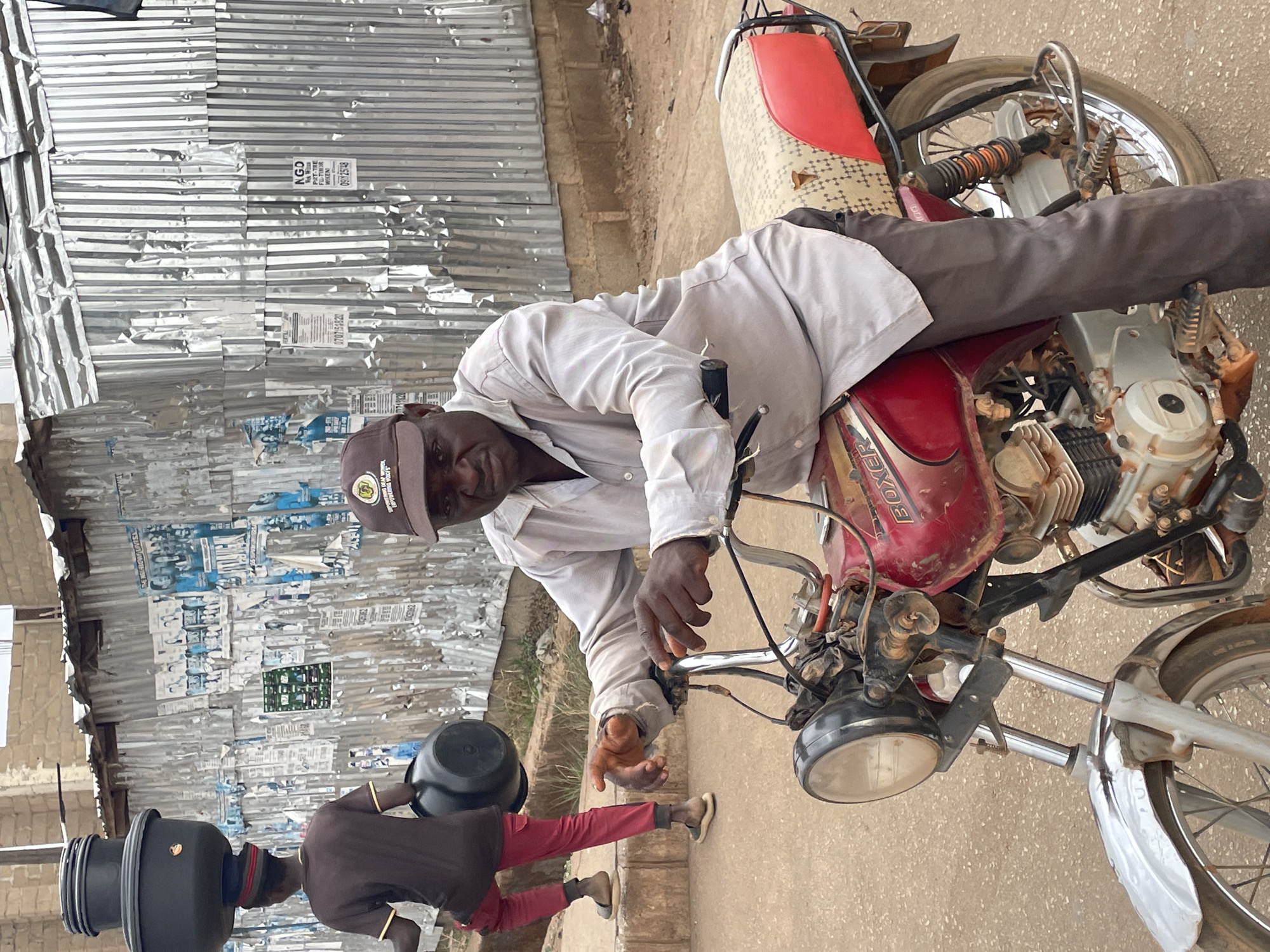 A man wearing a light-colored, long-sleeved shirt, grey trousers, and a brown cap with a circular emblem sits astride a red Boxer motorcycle, holding the handlebars. Another man in red trousers and a brown shirt is standing near a bucket, holding another bucket. Behind them, a corrugated metal wall displays torn posters and graffiti. Some posters display the following text "NGD Rok Wisdom", "PIZT-TRE", "FULL TIREOR", "091125148", and "0707151822". The scene appears to be in an outdoor location. The city is presumed to be Jos, Nigeria.