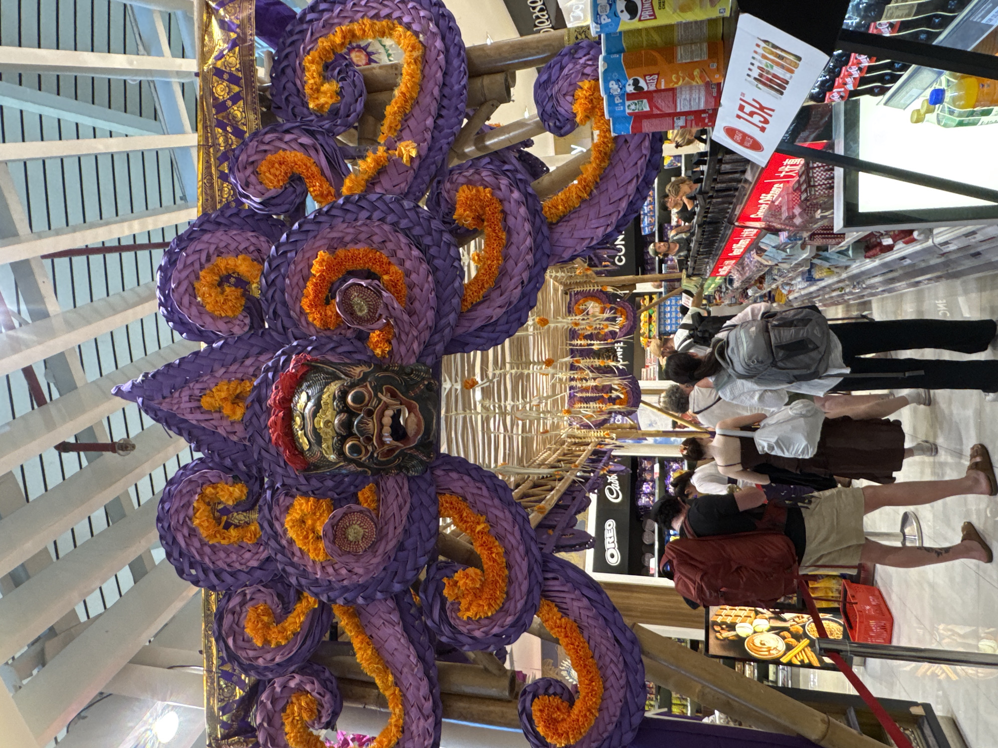 The photograph captures an indoor scene in Kuta, Indonesia, featuring a prominent artistic structure adorned with purple and orange floral and woven decorations. The structure includes a central dark-colored mask with detailed facial features.
People are present in the scene, with at least five individuals visible. One person wears a black t-shirt, khaki shorts, and has a red backpack, while another is dressed in a white shirt and dark skirt. Shoppers can be seen browsing in the background.
The environment appears to be a commercial or retail space, evident from the presence of shelving units stocked with products such as snacks and drinks. A price tag with the number "15k" is visible, and the brand "OREO" can be distinguished on one of the shelves. There are other identifiable objects, including shopping baskets. The lighting is typical of an indoor commercial setting, contributing to the overall clarity of the image.