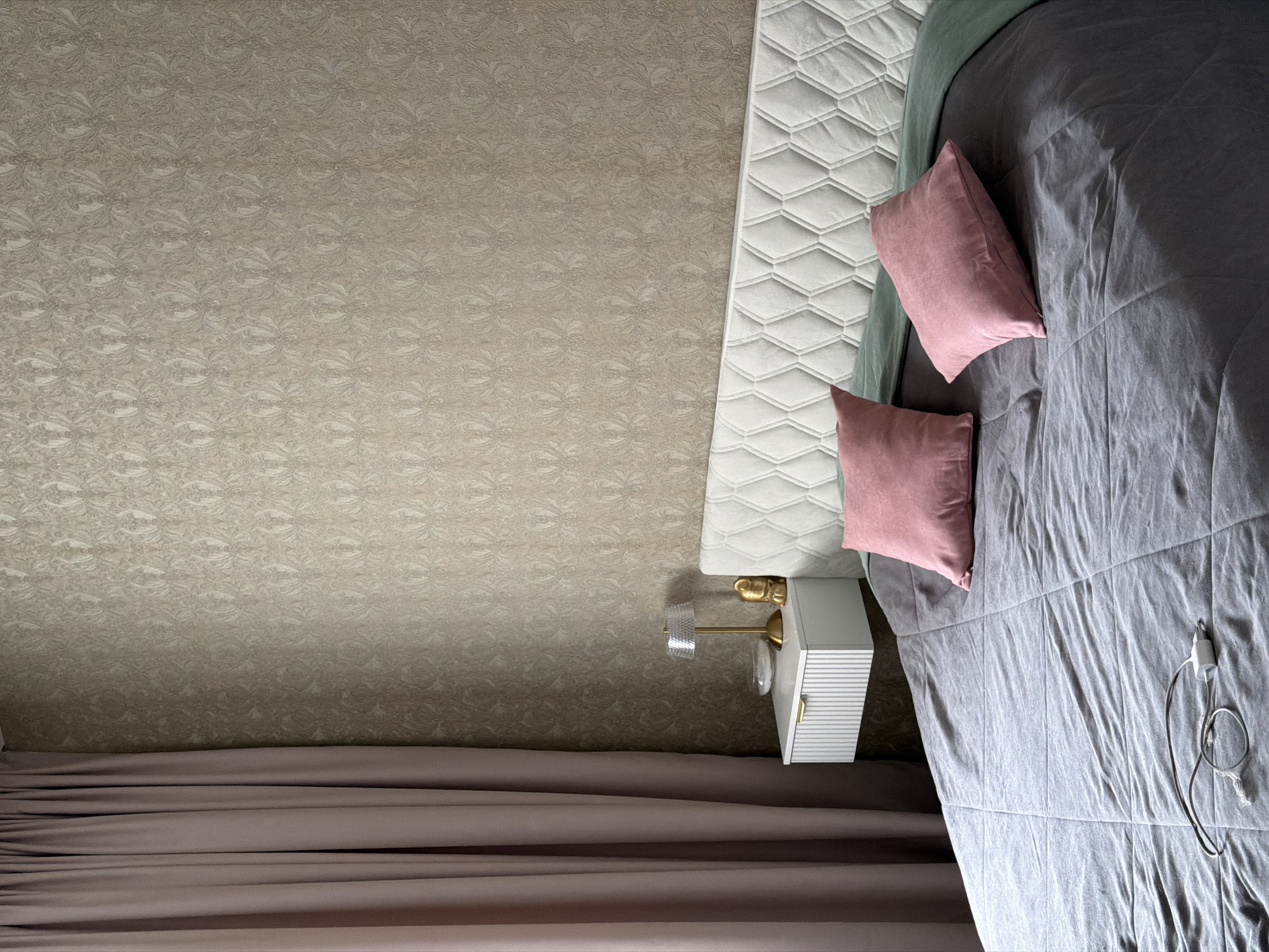 A low-angle, high-angle indoor shot of a bedroom, seemingly in Penza, Russia, features a neutral color scheme with accents of pink and gold. The image is oriented with the ceiling wallpaper dominating the upper portion, descending into a partially visible bed and bedside table in the lower right quadrant. A mauve curtain is visible at the bottom edge. The wallpaper has a muted floral pattern. The bed is covered in a gray duvet, with two pink square pillows. A white nightstand with a gold handle and a clear, decorative lamp sits beside the bed. A charging cord with a white plug is visible on the bed. No people are present in the image.
