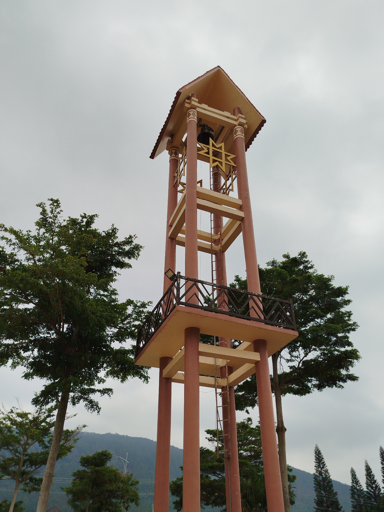 The image features a tall, four-tiered bell tower in K'Long, Vietnam. The tower is constructed with red-brown support columns and tan-colored platforms and roof structure. A ladder is affixed to one side of the tower, providing access to the upper levels. At the top of the tower, a large, dark-colored bell is visible, positioned behind a gold geometric design. The tower has a small viewing balcony with a railing. Two green trees flank the tower. The sky is overcast. In the background, a mountain range is visible.