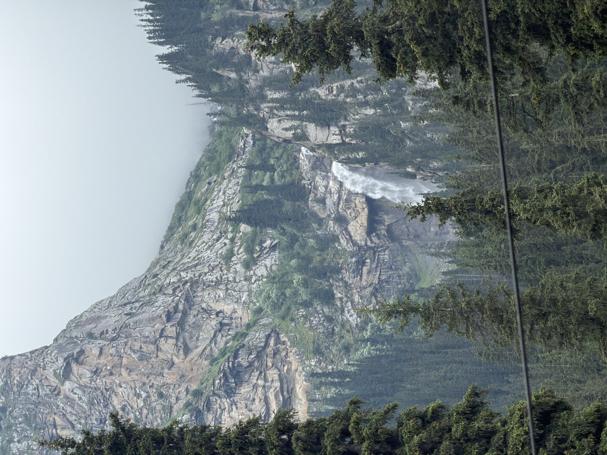 The image depicts a majestic mountain landscape with a prominent waterfall cascading down a rocky cliff face. Dense evergreen trees blanket the slopes leading up to the waterfall, their dark green foliage contrasting with the lighter grey and brown hues of the rock. The sky is overcast with a soft, diffused light, suggesting a cloudy day or early morning. In the foreground, the tops of more evergreen trees are visible, framing the scene. The overall impression is one of natural grandeur and a remote, wild environment, likely situated in a mountainous region of Kyrgyzstan. The specific location within the city of Unknown, Kyrgyzstan, is not determinable from the image alone. There are no people or visible human activity in the scene.