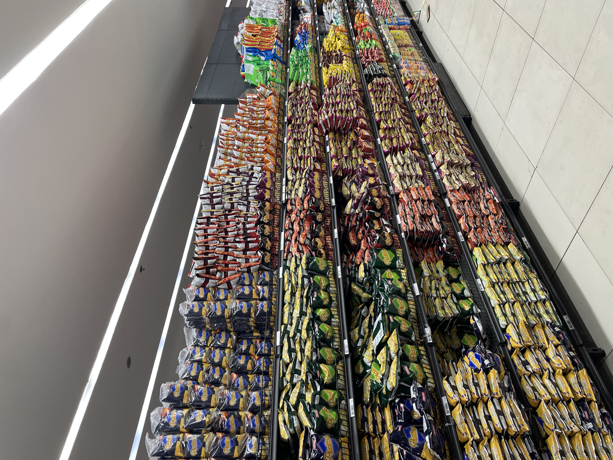 The image shows rows of snack food bags displayed on shelves in a supermarket aisle in Davao City, Philippines. The shelves are stocked with a variety of bagged snacks, predominantly chips, in different colors and packaging, suggesting a wide selection of flavors and brands. The lighting appears to be artificial, with fluorescent lights visible on the ceiling. The floor is tiled with large, light-colored square tiles. There are no people visible in the image. The overall scene is a well-organized retail environment.