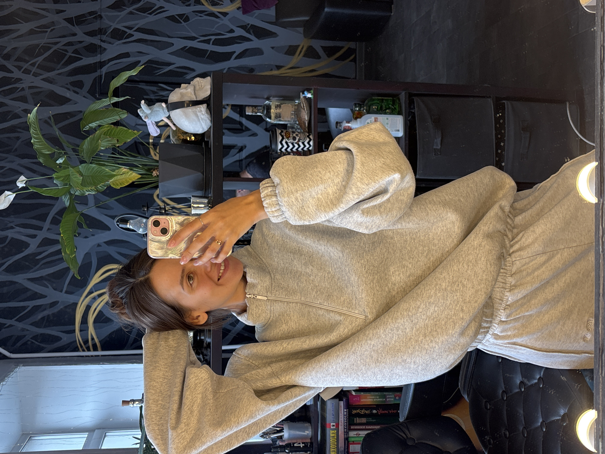 A person with brown hair is taking a selfie in front of a mirror. The person is wearing a gray zip-up sweatshirt and gray sweatpants. The background includes a plant, shelves with various objects, a black chair, and a wall with a dark, abstract pattern. The location is Krasnoyarsk, Russia.