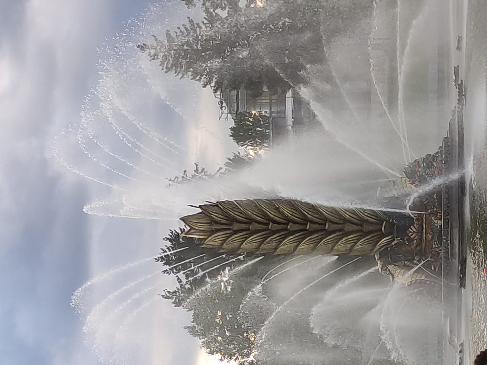 Here is a detailed description of the image:
The image showcases a large, elaborate fountain in an unidentified urban area. The fountain's centerpiece features a tall, sculpted structure resembling a golden wheat stalk or a similar elongated organic form. Water jets shoot outwards from the sides of this central structure in graceful arcs, creating a visually stunning display. The lower portion of the fountain appears to have ornate detailing and possibly depictions of figures or patterns. The background consists of trees and buildings, somewhat obscured by the spray of the fountain. The sky is partly cloudy. The overall impression is one of a public space with a significant water feature, likely intended to be a landmark or focal point within the city.