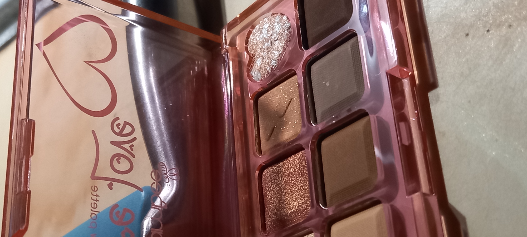 This close-up, angled shot features an open eyeshadow palette with a warm, neutral color scheme. The palette is housed in a transparent or translucent rose-gold colored plastic casing. The lid, partially visible on the left side of the frame, displays a large, stylized heart graphic in a darker pink hue. Below the heart, some text is visible, appearing upside-down and mirrored, which includes the letters "SVGA" and the partial word "palette" (specifically "ette"), along with a small, intricate bee or butterfly graphic.
The right side of the image focuses on the eyeshadow pans. There are several square-shaped pans containing various shades of brown and beige eyeshadow, ranging from light to medium-dark tones. At the top left of the eyeshadow section, a distinct heart-shaped pan is filled with sparkling rose-gold glitter. One of the square brown eyeshadows has a unique lightning bolt design embossed into its surface. The inner surface of the lid shows some reflections and minor smudges. The background appears to be a light-colored, slightly textured surface, suggesting an indoor setting. No individuals are present in the image.