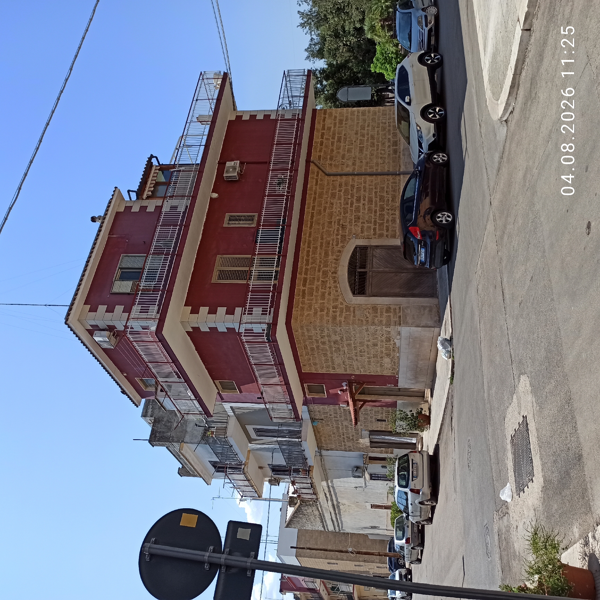 This is a street-level photograph taken in Bitonto, Italy, on August 4, 2026, at 11:25 AM. The scene depicts a residential building with a distinctive red facade on the upper stories and a rough stone base on the lower level. The building features multiple balconies with white railings. Several cars are parked on the street, including a dark blue hatchback, a white crossover SUV, and a silver sedan. A traffic sign is visible on the left side of the image, partially obscuring the view of the street and the building. The sky is clear and blue, and trees are present in the background on the right side of the image. The street is paved with asphalt and shows some wear and tear, including a manhole cover with debris on it. A white plastic bag is also on the street near the curb. The overall impression is of a quiet, sunny day in a residential area.