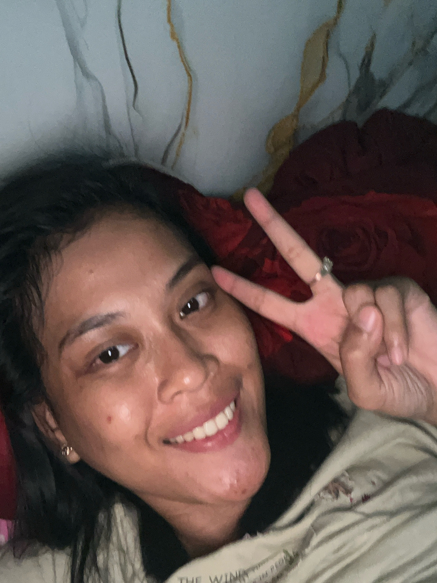 An overhead selfie shows a young adult woman with dark hair and brown skin lying on a bed. She is looking directly at the camera with a slight smile. Her right hand is raised and forming a peace sign. She is wearing a light-colored top and a ring on her right ring finger. The background is out of focus, but appears to be a patterned wall and a red blanket. The image was taken indoors in what is presumed to be a bedroom. No specific location in Kecamatan Pati, Indonesia, can be determined from the image.