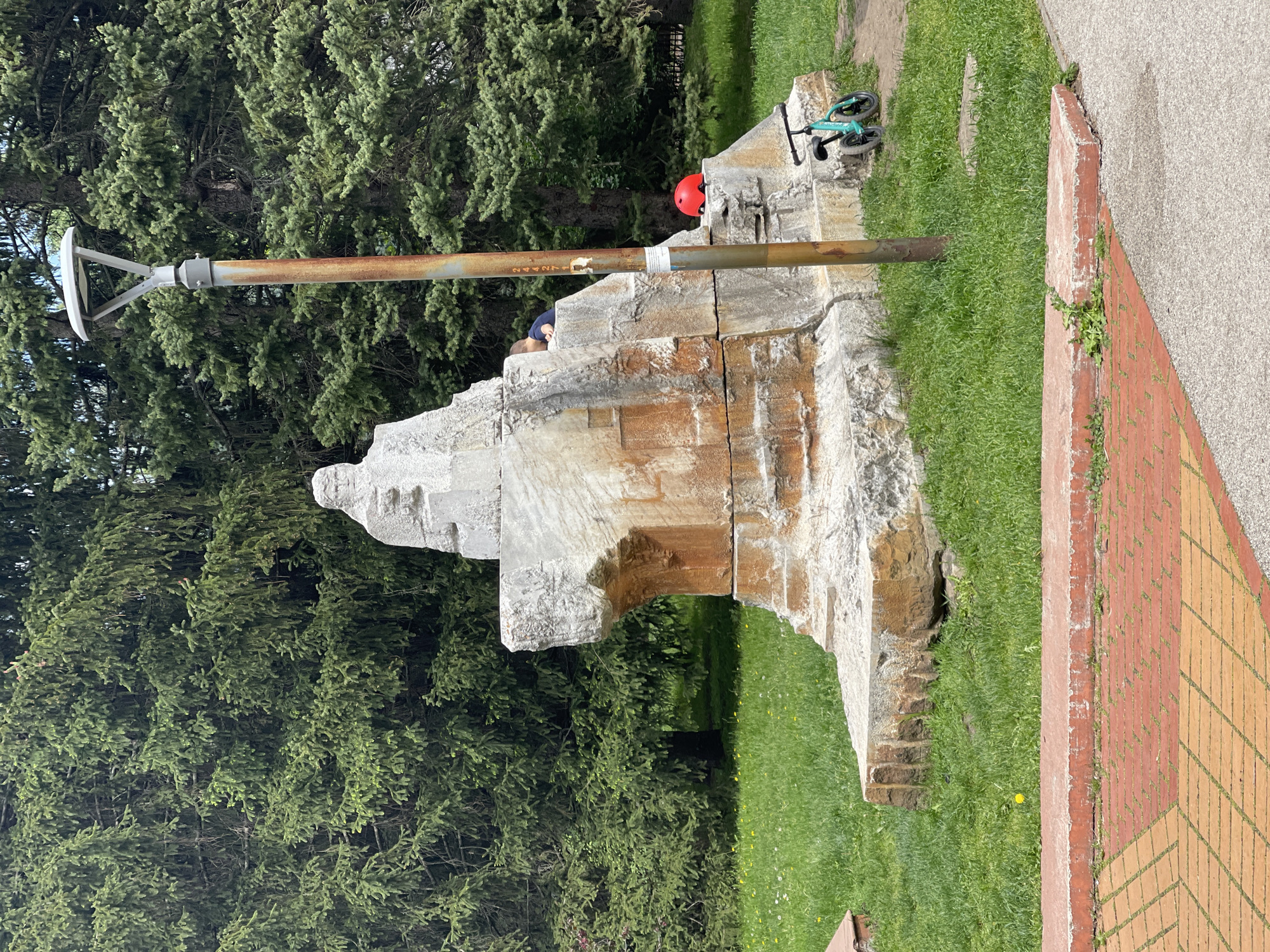 The image depicts an outdoor scene, likely in a park setting within the city of Sofia, Bulgaria. The focal point is a large stone sculpture or monument, constructed from rough-hewn blocks, possibly of marble or limestone. The monument has stepped levels and a somewhat abstract form. A rusty metal pole is affixed to the monument. A white lamp or sensor is attached to the pole. To the right of the monument, there is a brick pathway. A child's red helmet, and a small turquoise balance bike rest on the monument. The surrounding environment consists of green grass and coniferous trees. A child is visible sitting on the monument, under the helmet.