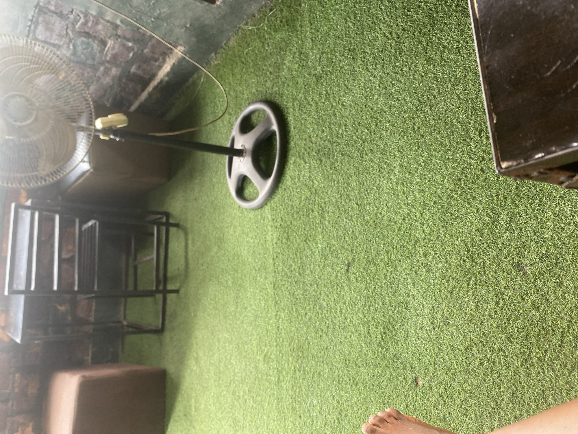 A personal perspective shot captures an outdoor setting with artificial turf flooring. In the foreground, a bare foot is visible, suggesting a relaxed atmosphere. To the left, a standing fan with a silver grill is angled, its stand extending towards the center of the frame. Behind the fan, a dark, rectangular object is partially visible, possibly furniture or a storage unit. Further back, a set of black metal shelves or racks stands, with some indistinct items stacked on them. The background features a brick wall on the left and a dark, flat surface on the right, which could be a table or counter. The overall scene appears to be an outdoor seating area or a recreational space, possibly a patio or a rooftop, with artificial grass laid out. The lighting suggests it could be daytime, with no strong indications of specific weather conditions. The image lacks any visible text.