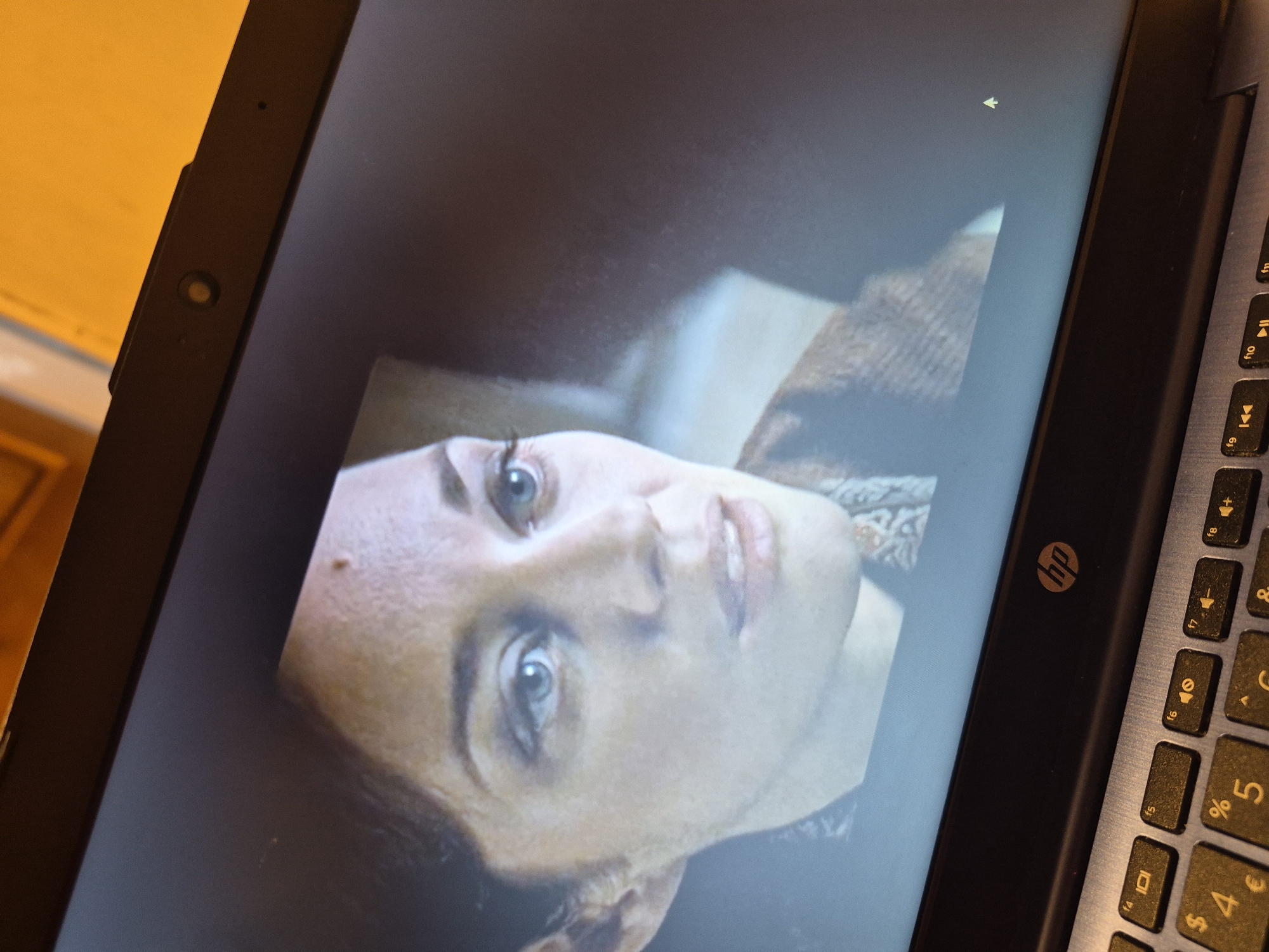 An extreme close-up shot is focused on a laptop screen displaying a scene from a movie. A woman's face fills most of the screen, with her blue eyes looking slightly off-camera to the right. Her hair is dark brown and pulled back, and she has a small mole on her forehead. She is wearing a textured, earth-toned garment, possibly a sweater or shawl. The background on the screen is dark and out of focus, with a suggestion of light coming from the right. The laptop itself is visible at the bottom of the frame, showing a portion of the keyboard and the HP logo. The lighting in the room appears to be warm and ambient, casting a slight orange hue on the top edge of the laptop. There is no discernible indication of the city of Sandwell, United Kingdom, within the image.