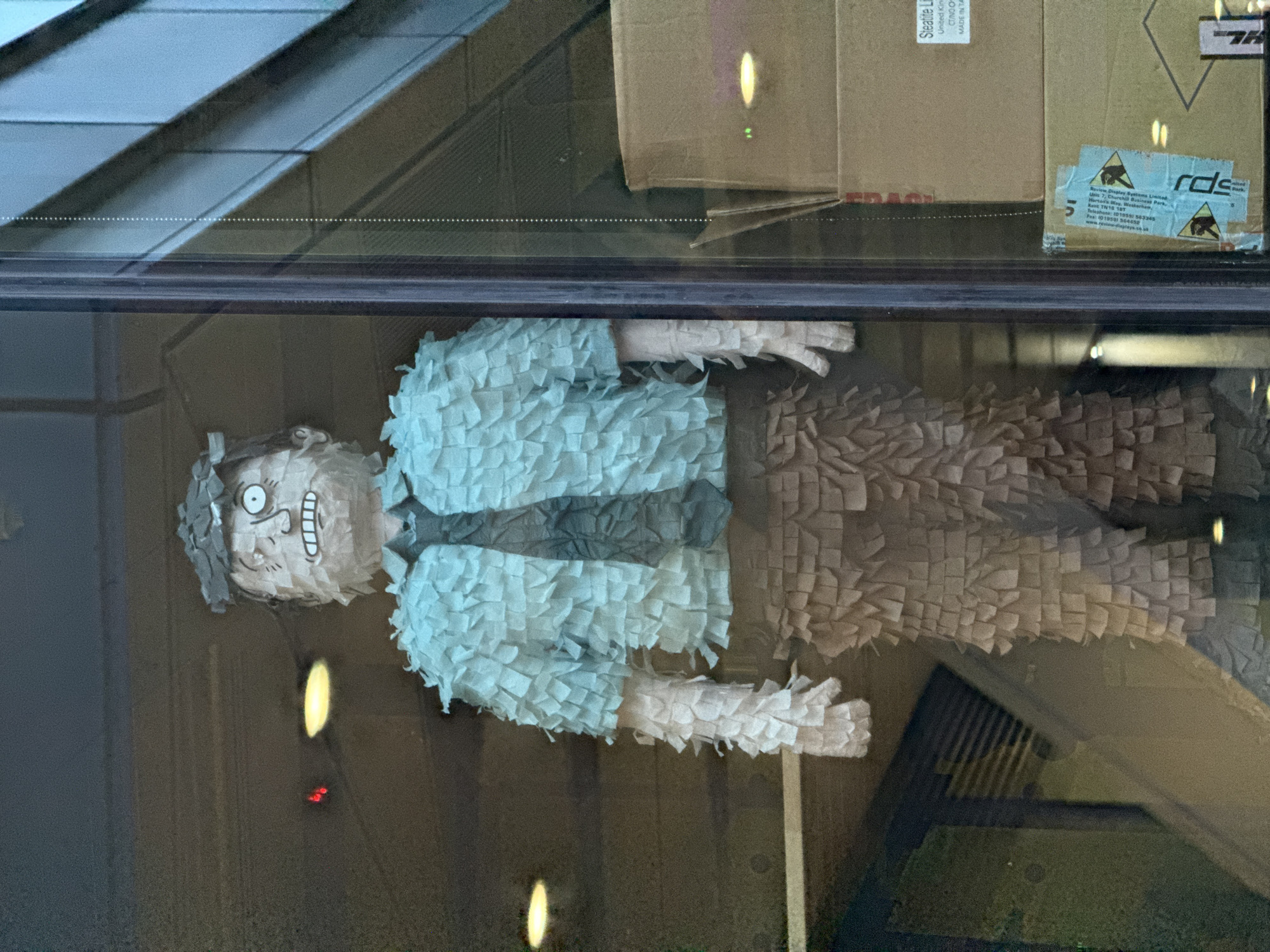 The image shows a homemade piñata in the likeness of a man, presumably displayed in a window. It has a papier-mâché head and a body covered in fringed paper, with a light blue shirt, gray tie, and brown pants. The piñata is indoors, positioned against the window, with reflections of the outdoors and interior lights visible on the glass. Above the piñata, partially visible in the window reflection, are cardboard boxes with labels. One label is marked with the words "Steatite Limited" and "MADE IN TAIWAN". Another is a caution label with the text "Review Display Systems Limited," and an address in Westerham, Kent, TN16 1BT, England. Based on the address, it seems unlikely that the image was taken in London.