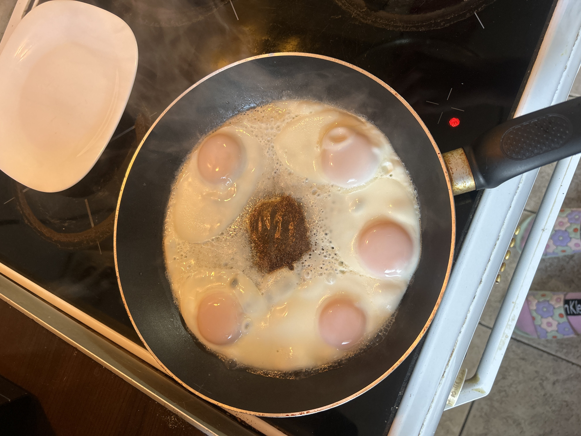 A top-down shot reveals a cooking scene, likely in a home kitchen. The focal point is a black frying pan on a black stovetop. The pan contains five fried eggs arranged in a circular pattern around a central mass of what appears to be burnt food or spices. Steam rises from the pan, indicating recent or ongoing cooking. To the left of the pan sits a white, square plate. A stove handle is visible in the upper right, and a portion of the person standing is visible on the side wearing flower decorated slippers. The image suggests the scene may be in Sofia, Bulgaria. The background shows parts of the stovetop and nearby kitchen elements, indicating the setting. The overall composition implies a common domestic activity, cooking.