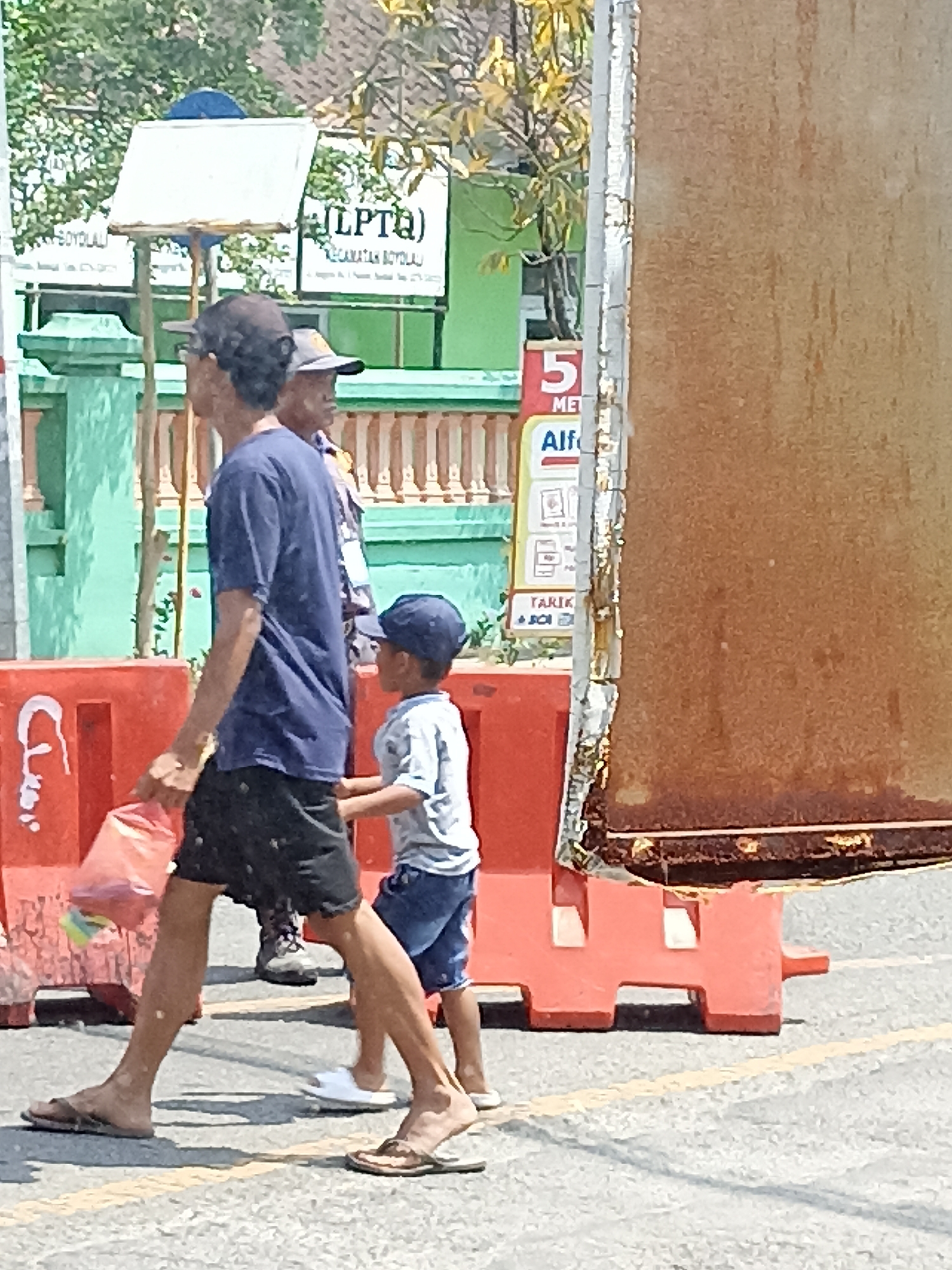 A man wearing a dark blue t-shirt and black shorts walks on a paved street, carrying a pink plastic bag in his right hand. He is wearing brown flip-flops. Beside him, a young boy in a light blue t-shirt and blue shorts, wearing a dark blue baseball cap, walks with his right hand on the man's left leg. Behind them, another person, partially obscured, is visible, wearing a hat and a patterned shirt. In the background, there are several signs, including one that reads "(LPTQ)" with "KECAMATAN BOYOLALI" below it. Red plastic barriers are lined up on the side of the street. The scene appears to be in Pulisen, Indonesia, suggested by the signage.