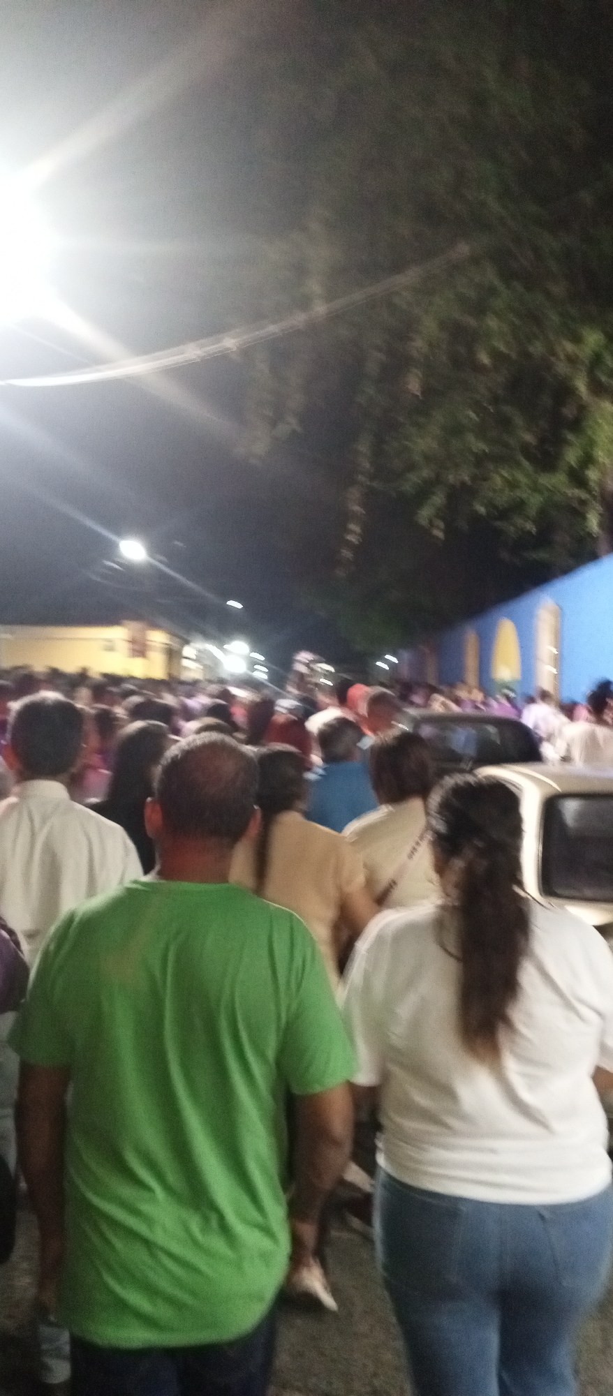 A crowd of people walk along a street at night. Most of the people in the foreground are facing away from the viewer. A man in a green shirt and a woman in a white shirt and blue jeans are walking towards the center of the crowd. A building with a blue facade and arched windows is visible on the right. A bright light is visible in the upper left corner of the image. The event takes place in the city of Tinaquillo, Venezuela.