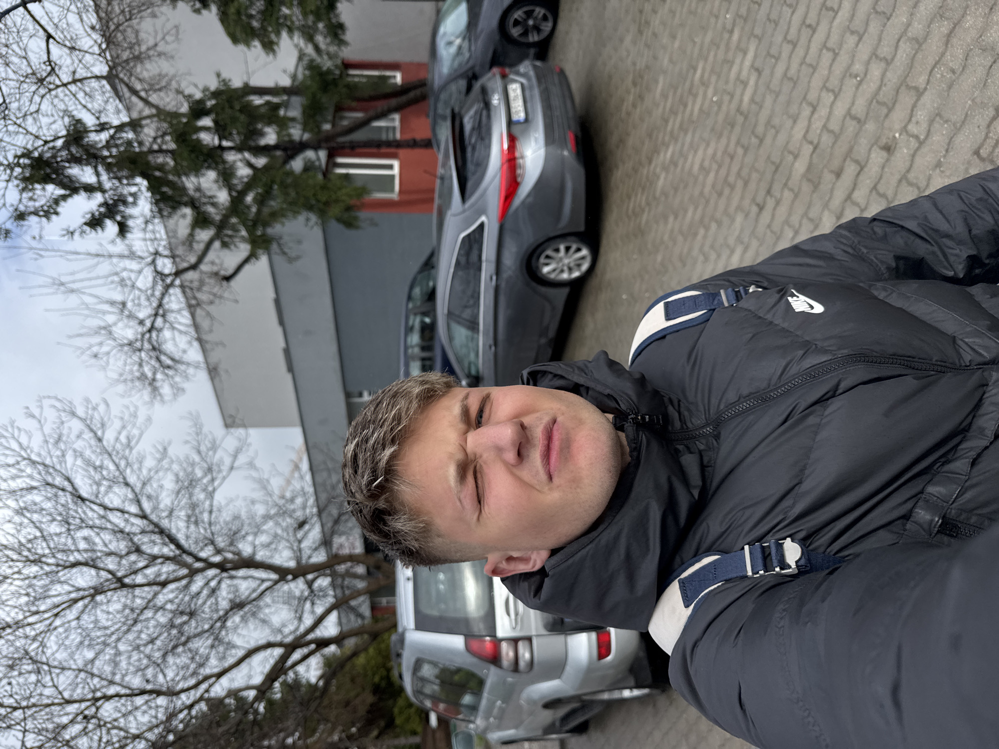 A man is taking a selfie outdoors. He is wearing a black Nike puffer jacket with blue and white straps. Behind him are parked cars on a paved surface, a building with a red section, and trees. The image suggests an overcast day. It is not possible to identify the exact location in Plovdiv, Bulgaria, without additional information.