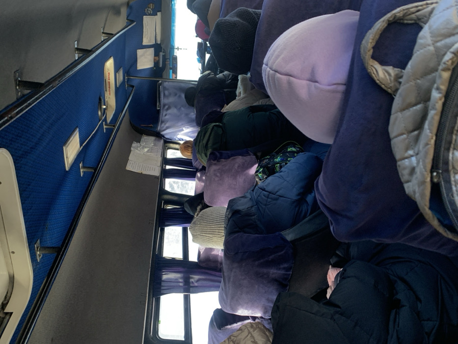 The scene depicts the interior of a passenger bus, observed from a forward-facing perspective, likely within the city of Kozyn, Ukraine. The bus is occupied by numerous individuals, predominantly visible from behind, seated in rows of dark blue or purple fabric seats. Many seats feature additional lighter purple or lavender cushions.
The overhead area features a blue, patterned ceiling with two rectangular white light fixtures and several structural supports. Grey side panels run along the upper wall sections above the windows. Blue curtains are drawn on the windows, through which a bright, featureless exterior, possibly snow-covered ground or overcast sky, is visible. A white sign with red text, possibly indicating an emergency exit, is mounted on the ceiling, along with other papers or documents taped to an overhead panel. A small cross pendant hangs near the front windshield area.
Passengers are dressed in winter attire, including quilted jackets in shades of blue, dark green, and black, and knit hats in light beige and dark grey. Personal belongings such as a black and green patterned backpack and a light grey quilted backpack are visible in the foreground and among the passengers. No direct interactions between individuals are discernible. All visible individuals are seated, occupying the majority of the available seating.
