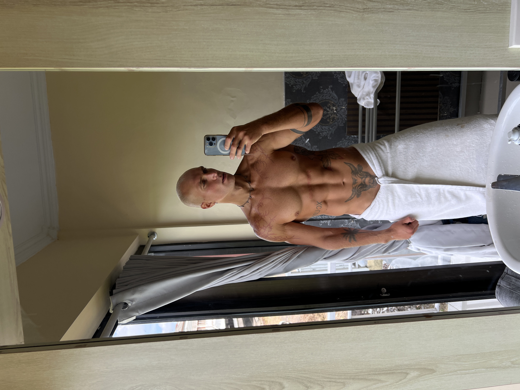 A bald, tattooed man is taking a mirror selfie in what appears to be a bathroom in North Kuta, Indonesia. He is wearing a white towel wrapped around his waist, exposing his toned torso and abdomen. The man is holding a smartphone in his right hand, pointed at the mirror. In the background, a window with gray curtains is visible, offering a glimpse of the outdoors. The bathroom features wooden paneling on the right side and a patterned wallpaper on the back wall. A sink and faucet are partially visible in the foreground.