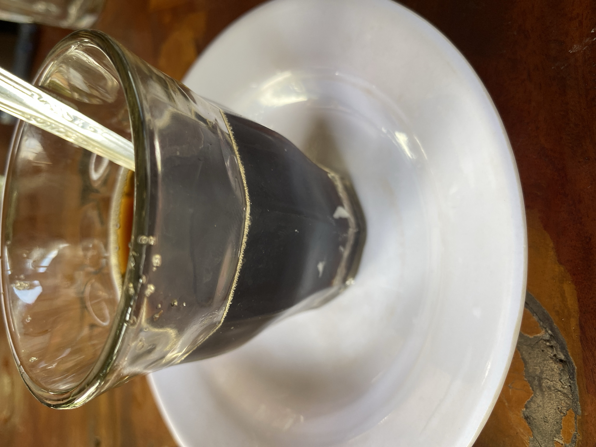 A close-up, overhead shot shows a clear glass of dark coffee with a small spoon inside. The glass is resting on a white saucer, which is on a wooden table. The coffee has a frothy layer on top, and small bubbles are visible on the inside of the glass. The spoon is silver and has a decorative pattern on its handle. The wooden table has a textured surface with variations in color, suggesting it is aged or made of a natural wood. The lighting in the image is soft, with reflections visible on the glass and saucer. The image appears to be taken in Aluepiet, Indonesia, likely in a cafe or dining setting.