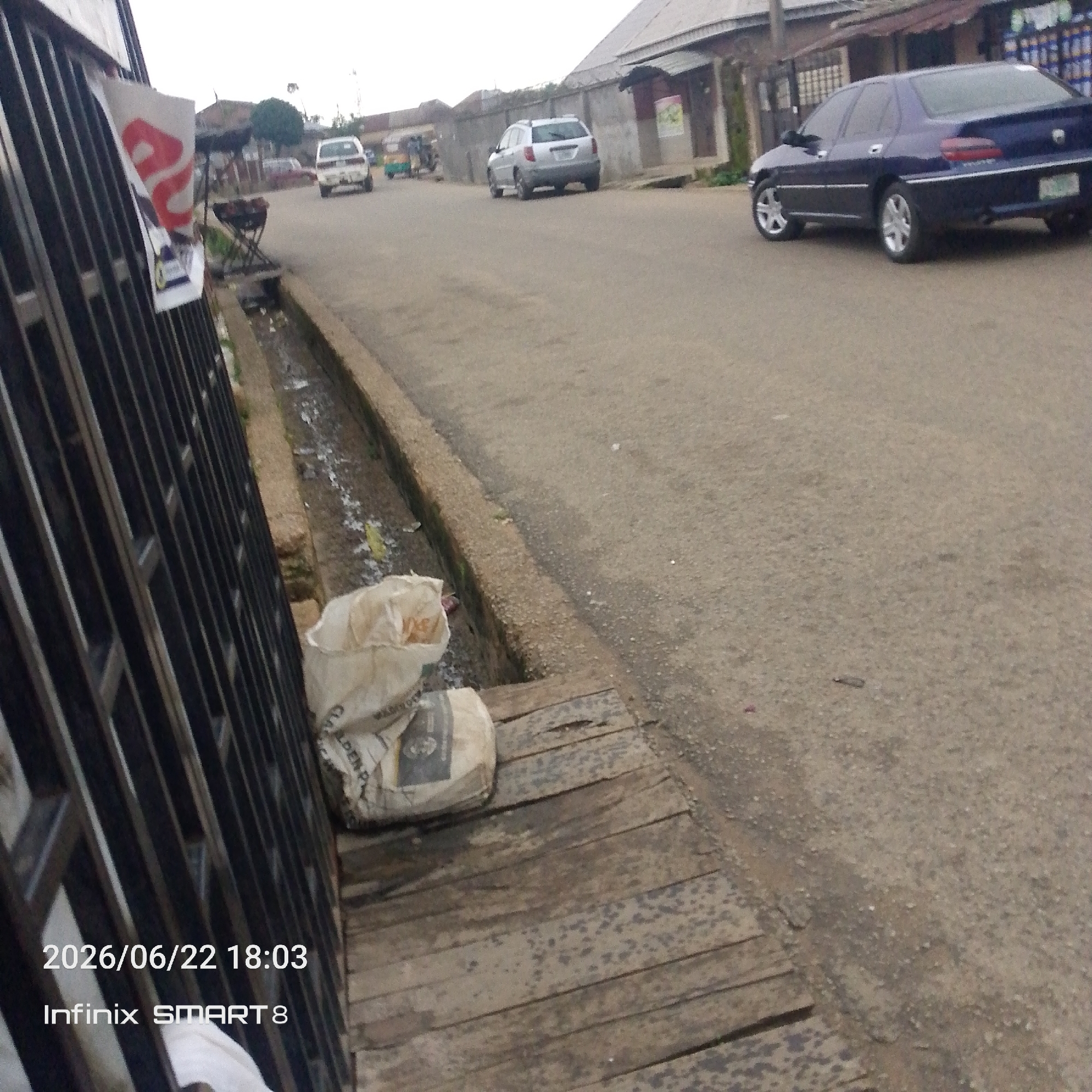 The image depicts a street scene in Jos, Nigeria, likely during daylight hours given the brightness. In the foreground, a black metal gate is visible on the left, with a white poster attached to it. Two large bags, appearing to be filled with powder, are placed on a wooden structure next to the gate. One bag is partially unzipped, revealing white contents. A shallow concrete drainage channel runs alongside the street.
The street itself is paved and appears to be a moderately busy area. Several vehicles are present: a dark blue sedan is parked on the right side of the road, a light-colored hatchback is further up the street on the left, and a white car is also visible in the distance. A green and yellow auto-rickshaw or "Keke Napep" is also seen in the middle ground, suggesting local transportation. Buildings line the street, with a mix of residential and commercial structures.
The overall mood is one of everyday life in an urban environment. The sky is overcast, indicating possibly cloudy weather. Visible text includes a date and time stamp "2026/06/22 18:03" and "Infinix SMART 8" at the bottom left corner of the image, which are likely from the camera or phone that captured the photo and not part of the scene itself. There is also some text on the white poster on the gate, but it is not clearly legible. The bags also have some printed text, but it is too blurry to read.