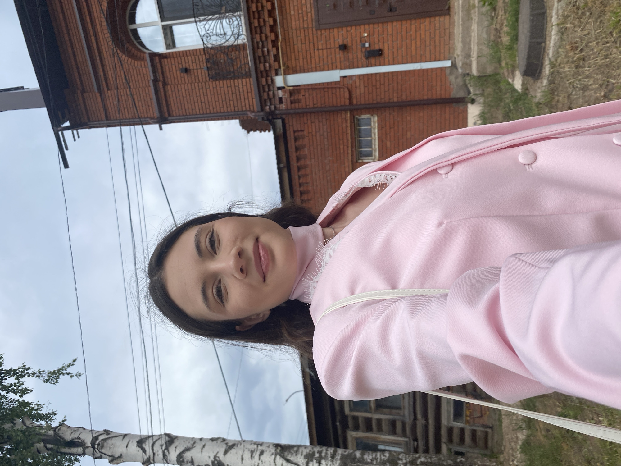 A young woman stands in Votkinsk, Russia, wearing a pink jacket, posing for a photo. Behind her are two buildings, one made of red brick with a curved window on the top floor, and another wooden structure. A birch tree and overhead power lines are visible on the left side of the frame, set against a cloudy sky.