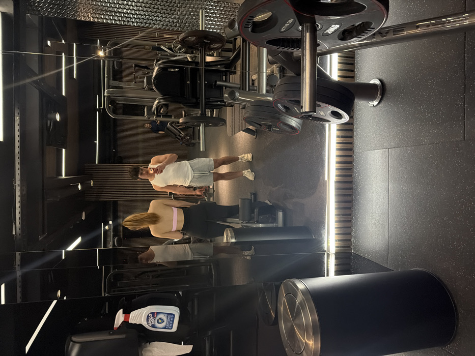 The image displays the interior of a modern gym located at ACTIVE GYM, Bulgaria Mall, Sofia, Bulgaria. The perspective is from the eye level of an individual holding a smartphone, whose reflection is prominent in the central mirrored wall. This male individual wears a white sleeveless top and grey shorts. To his left in the reflection, a female individual is visible from behind, facing away from the camera, with long reddish-blonde hair in a ponytail, wearing a light pink sports bra and black leggings. A third male individual in a dark t-shirt is visible in the background reflection on the far right.
The gym floor consists of dark grey speckled rubber matting. On the far left, a black cylindrical trash can with a brushed metallic lid stands beside the mirrored wall. Above it, a white "APOLO SEPT" branded disinfectant spray bottle is clipped to a dark stand, with a white paper towel dispenser below. Various gym equipment is scattered throughout the space, including weight machines, barbells, and 20kg weight plates branded "ACTIVE GYM" in the right foreground. The gym's interior design features dark, reflective floor-to-ceiling mirror panels, sections of vertical wooden slat wall, and a textured metallic wall segment on the far right. Lighting is provided by recessed ceiling spotlights and a continuous horizontal LED strip light illuminating the base of the mirror wall. The overall environment is clean, functional, and well-lit.