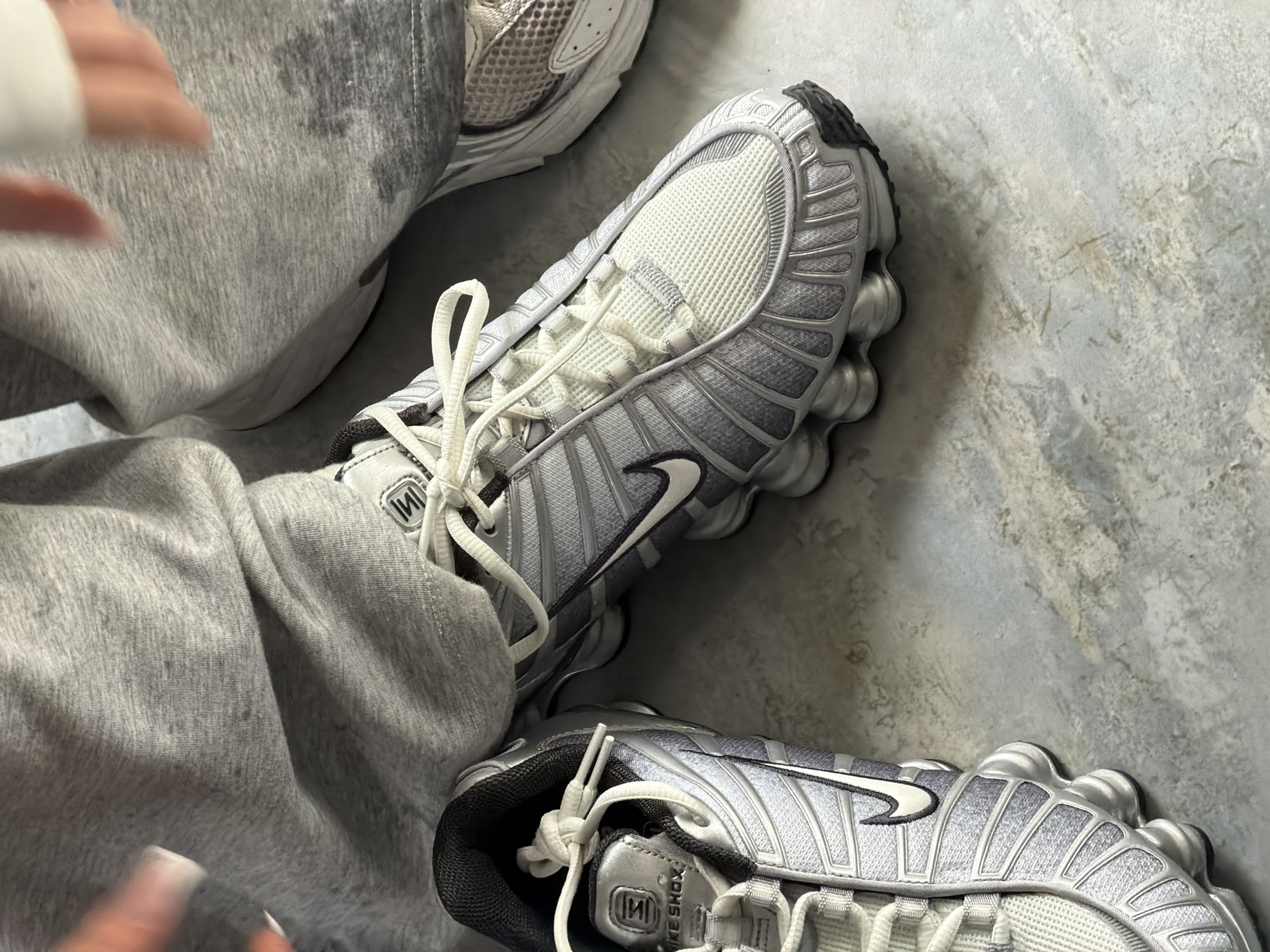 The image shows a pair of light grey Nike Shox TL sneakers on a person wearing grey sweatpants. The footwear is on a grey surface, possibly a floor, with another shoe partially visible in the upper left corner. One shoe is positioned closer to the viewer and the other angled away. The person’s fingers are visible on either side of the image, suggesting the picture was taken from a first-person perspective. There is no information to suggest that the location is Yerevan, Armenia.