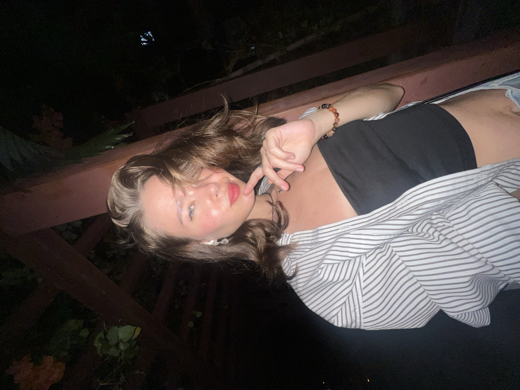 A young woman is posing outdoors at night. She is lying on a wooden structure, possibly a bench or low wall. She is wearing a black tank top, a striped open shirt, and what appears to be denim shorts. She has brown wavy hair and is wearing earrings and a bracelet. Her right hand is raised to her face, with her finger near her lips. The background is dark, with some foliage visible, and appears to be in a garden or park-like setting. The location is possibly within Nam Nha Trang Ward, Vietnam.