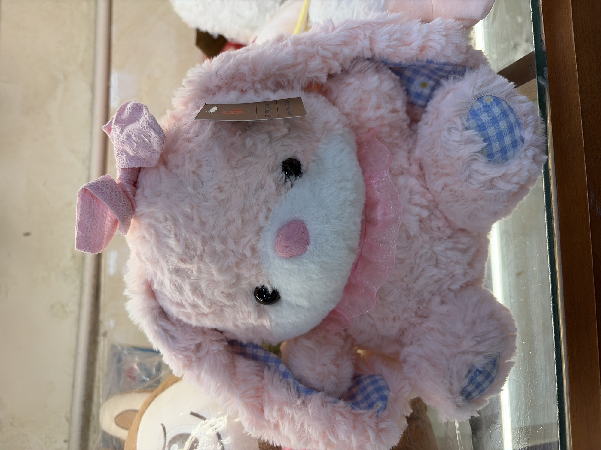 A pink, plush bunny stuffed animal is displayed on a glass shelf in what appears to be a retail store in Da Nang, Vietnam. The bunny has large, floppy ears lined with blue and white checkered fabric, and a pink bow on its head. It wears a ruffled pink collar around its neck. The bunny's paws also feature blue and white checkered fabric accents. A retail tag is attached to its ear. In the background, other stuffed animals and store merchandise are visible. The lighting suggests an indoor environment, possibly a toy store or a gift shop.