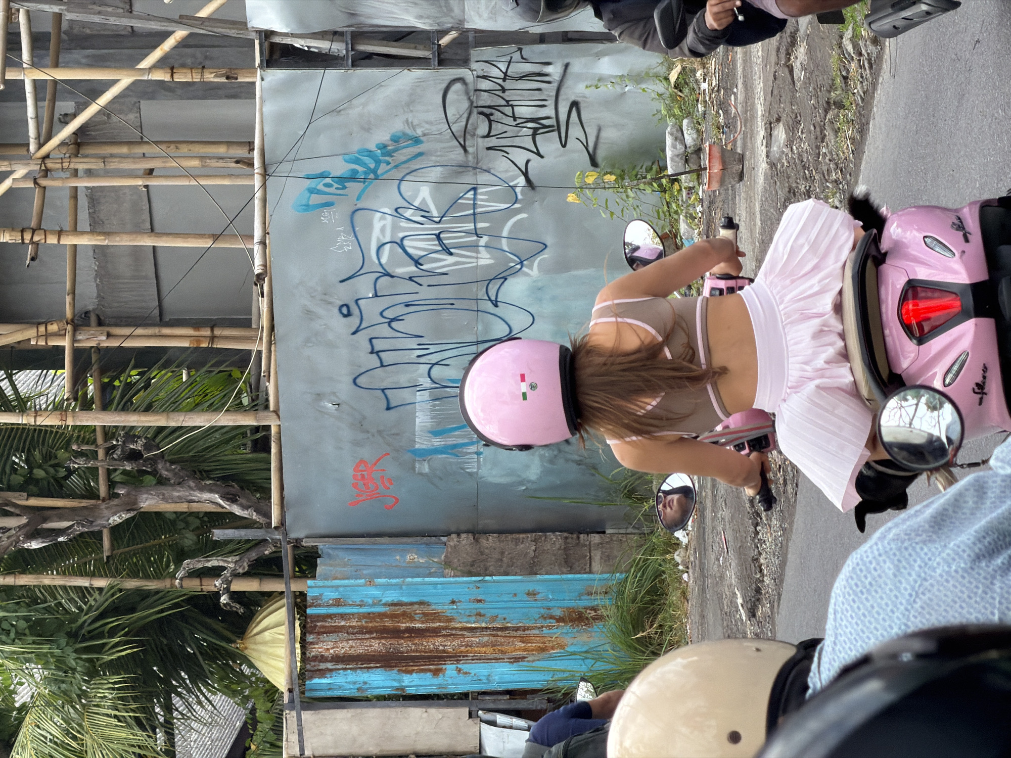 In North Kuta, Indonesia, two individuals are riding on a pink scooter. The person in the foreground, facing away from the camera, is wearing a pink helmet and a light-colored halter top with a pink trim, along with a pleated pink skirt. They are holding the scooter's handlebars. Behind them, another person, partially visible, is wearing a light blue patterned shirt and a beige helmet. The scooter is on a paved road with some debris and foliage on the side. A graffiti-covered wall is visible in the background, along with bamboo scaffolding and palm trees.