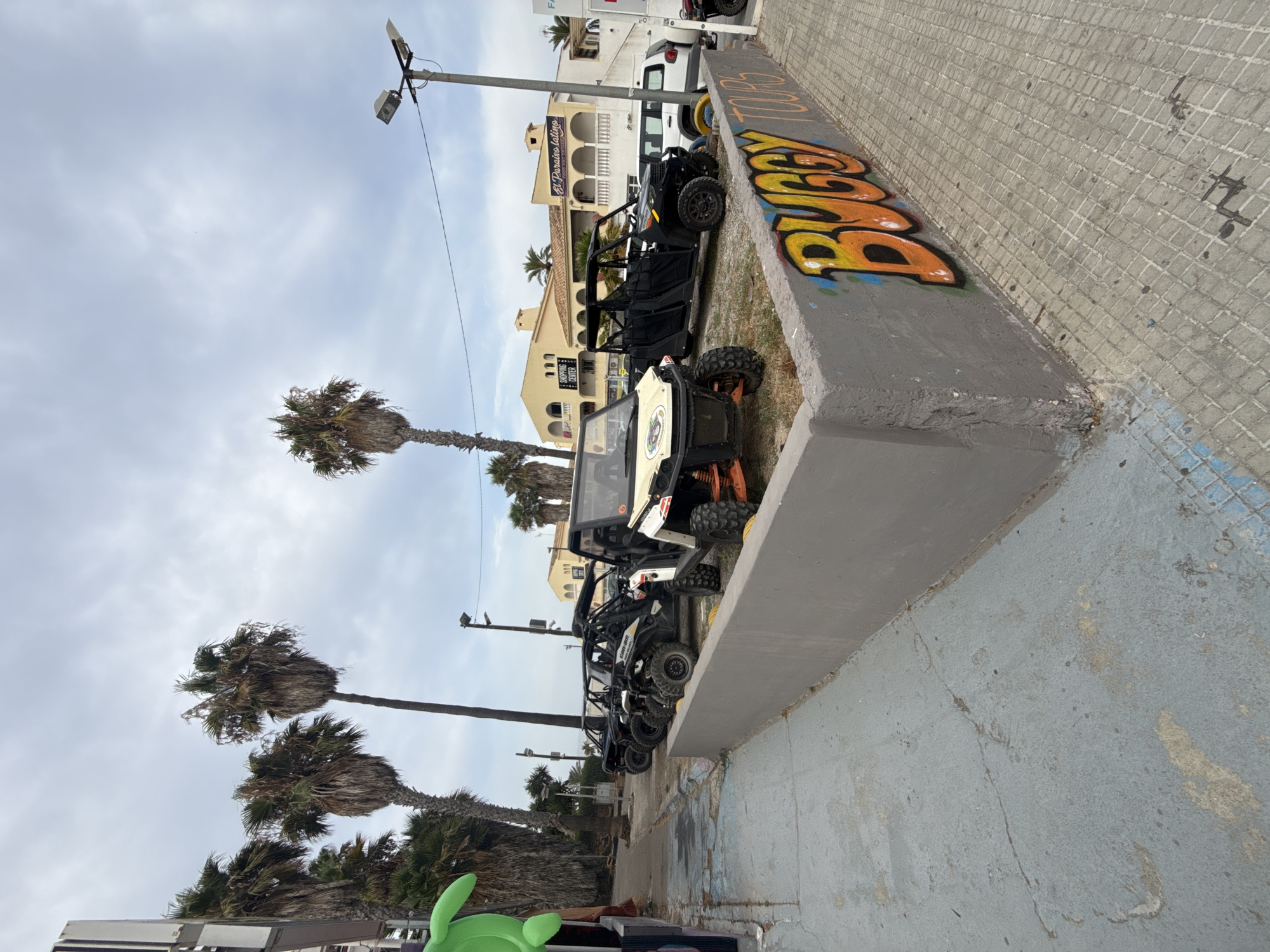 In Cales De Mallorca, Spain, a row of off-road buggies are parked on a paved surface. The buggies are of various colors, including black and white, and appear to be for rent or tour purposes. Some buggies have visible roll cages and high-clearance tires. In the background, there are palm trees, a light pole with multiple fixtures, and buildings, one of which has a sign that reads "SHOPPING CENTER." Another building has a sign that appears to say "El Paraiso Latino." Graffiti, including the word "BUGGY," is visible on a low concrete wall in the foreground. The sky is overcast.