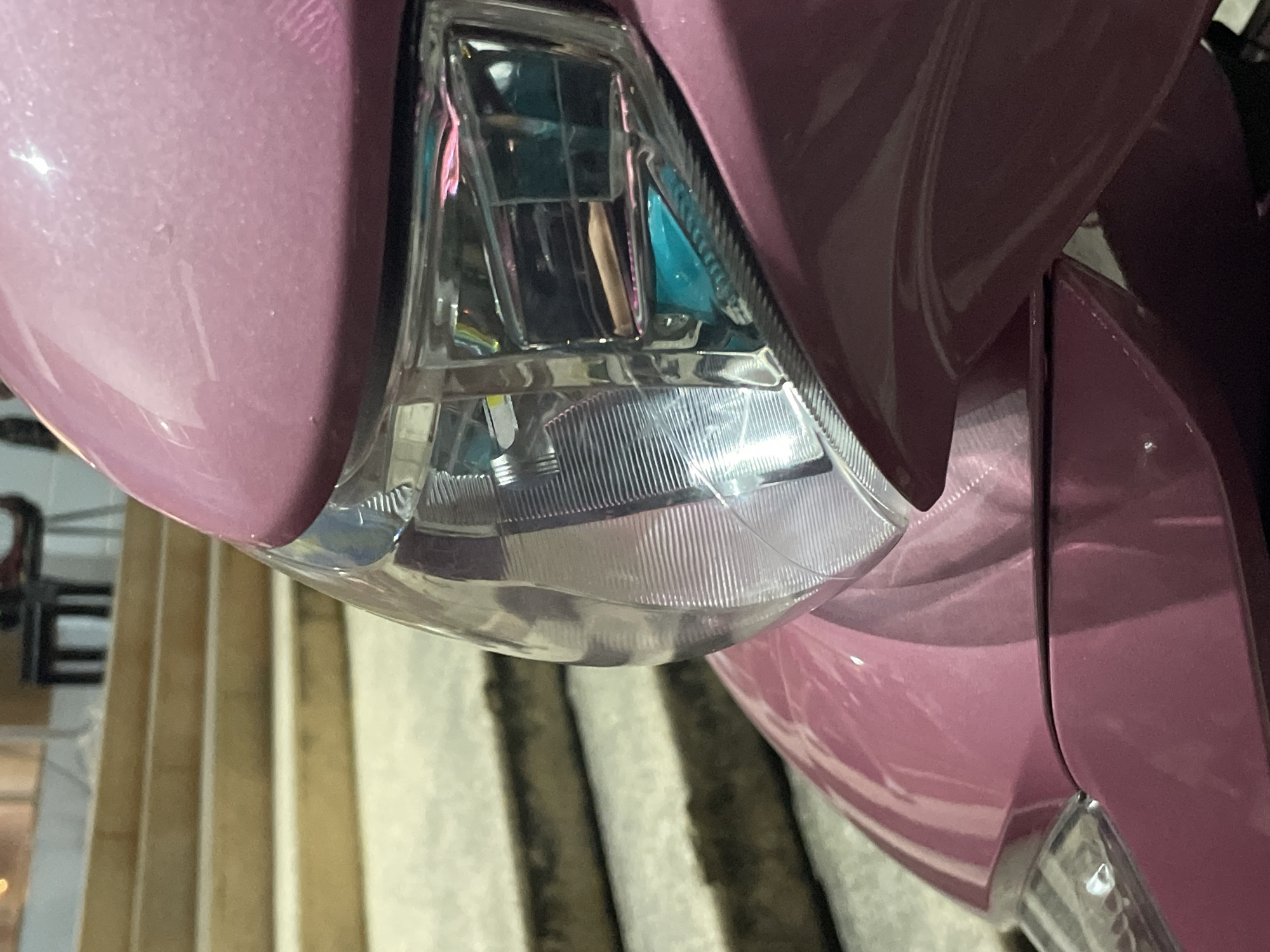 A close-up, eye-level shot focuses on the pink-painted front of a motorcycle, specifically the headlight and part of the fairing. The headlight is clear plastic with a ribbed interior, reflecting a greenish-blue light within its housing. Behind the motorcycle, a set of concrete stairs with wooden planks on each step recedes into the background. The environment appears to be outdoors, possibly in a public area or a residential setting. The lighting is dim, suggesting it might be evening or nighttime. The scene is devoid of people or any overt action.