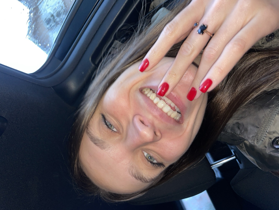 A smiling woman is seen from an unusual, tilted perspective, seemingly inside a car. The car's interior and a rain-streaked window are visible. She has fair skin, light eyes, and brown hair. Her teeth are prominent in the smile. Her fingernails are painted a vibrant red. A ring with a dark stone adorns one of her fingers. A dark car seat and the upper portion of a jacket are also visible.
