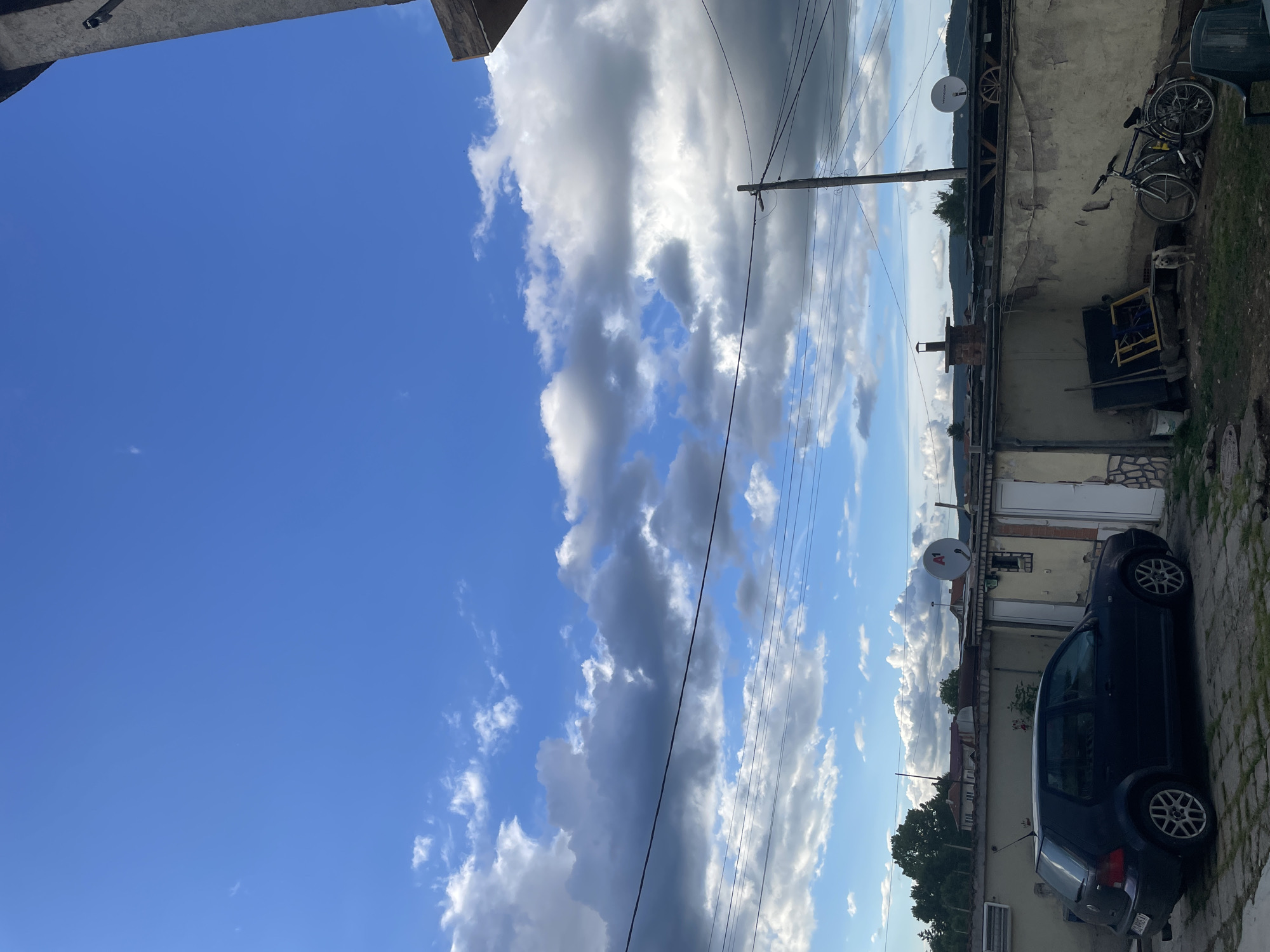The image, set in Ihtiman, Bulgaria, captures a sunlit outdoor scene under a vibrant blue sky with scattered, fluffy cumulus clouds. The sky occupies the upper portion, showcasing both bright white cloud formations and darker, shadowed areas, indicative of fair weather.
A prominent wooden utility pole with multiple overhead power lines cuts through the midground, extending horizontally across the frame. To the right, various single-story buildings or outbuildings line the scene. These structures exhibit a mix of light-colored, possibly stuccoed walls and some visible brickwork, suggesting an aged appearance. One building features a brick chimney, while another prominently displays two satellite dishes, one of which has "VIASAT" written on it. An old wooden cartwheel leans against one of these walls.
In the lower right, a dark-colored hatchback car, possibly a Volkswagen Golf, is parked on a paved surface that appears to be cobblestone or rough concrete. Its rear license plate, "CA 6917 MP," indicates a Sofia registration. Nearby, a blue bicycle is propped against a wall, and below it, a small, light-colored dog or statue of a dog rests on the ground amidst some miscellaneous clutter and wooden frames.
In the distance, faintly visible beneath the clouds, are hazy outlines of hills or mountains. The lighting suggests it is late morning or early afternoon, with clear sunshine. There is no specific activity taking place; the scene is static, depicting an ordinary moment in what appears to be a residential or backyard area.