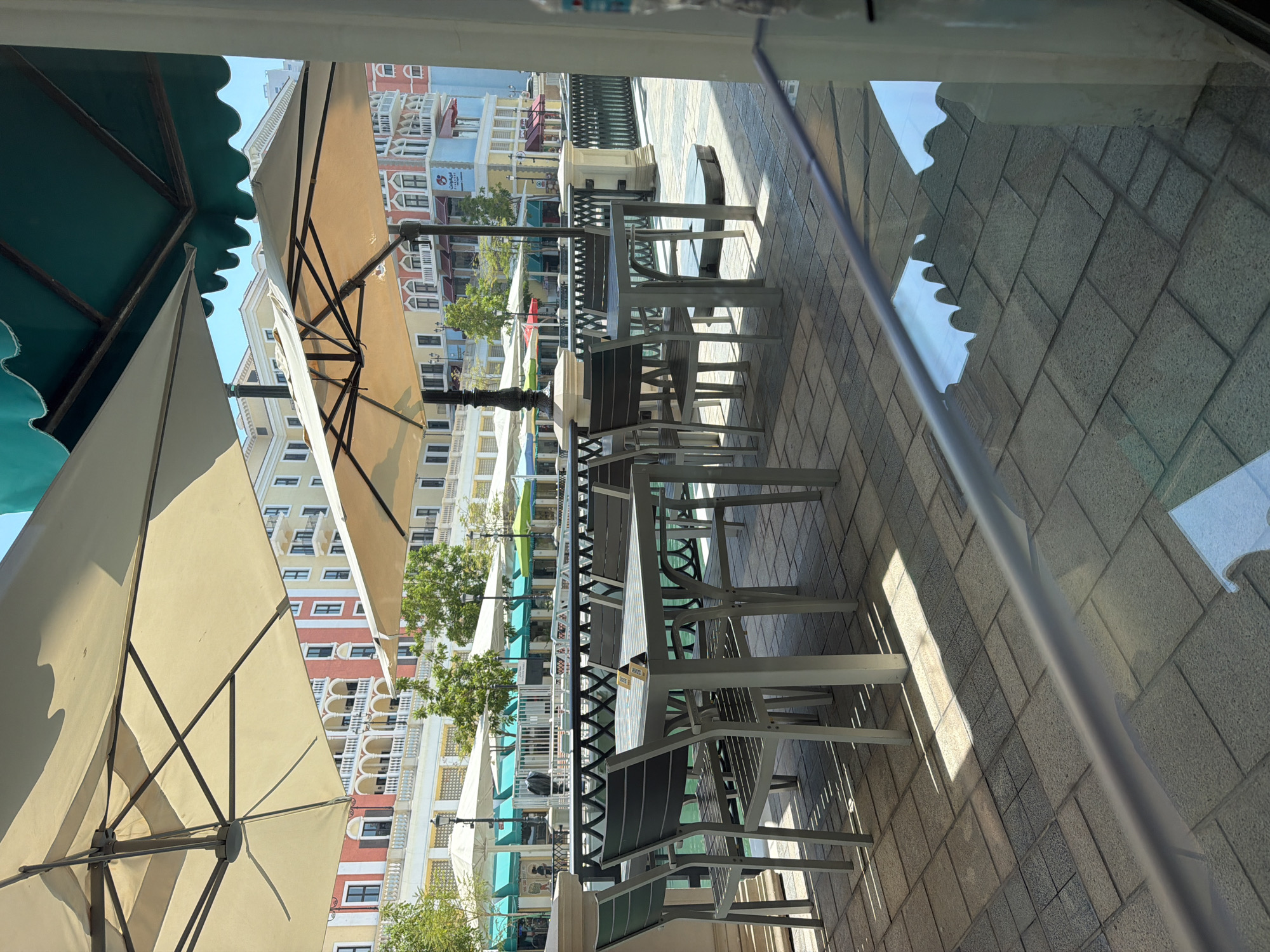 The image depicts an outdoor seating area at a waterfront establishment, likely a cafe or restaurant, in Doha, Qatar. The scene is characterized by a row of tables and chairs set on a tiled patio, shaded by large, light-colored umbrellas and a green awning. In the background, vibrantly colored buildings with architectural details reminiscent of Venetian or Mediterranean styles line the waterfront. Trees and smaller awnings of various colors are visible along the building facades. The water body in front of the buildings reflects the sky and the surrounding structures. The overall atmosphere suggests a pleasant, sunny day, with the seating area appearing ready for patrons, though no people are actively visible in the immediate foreground or midground. The perspective is from within a building looking outwards, with a glass barrier or window in the foreground creating reflections on the tiled floor.