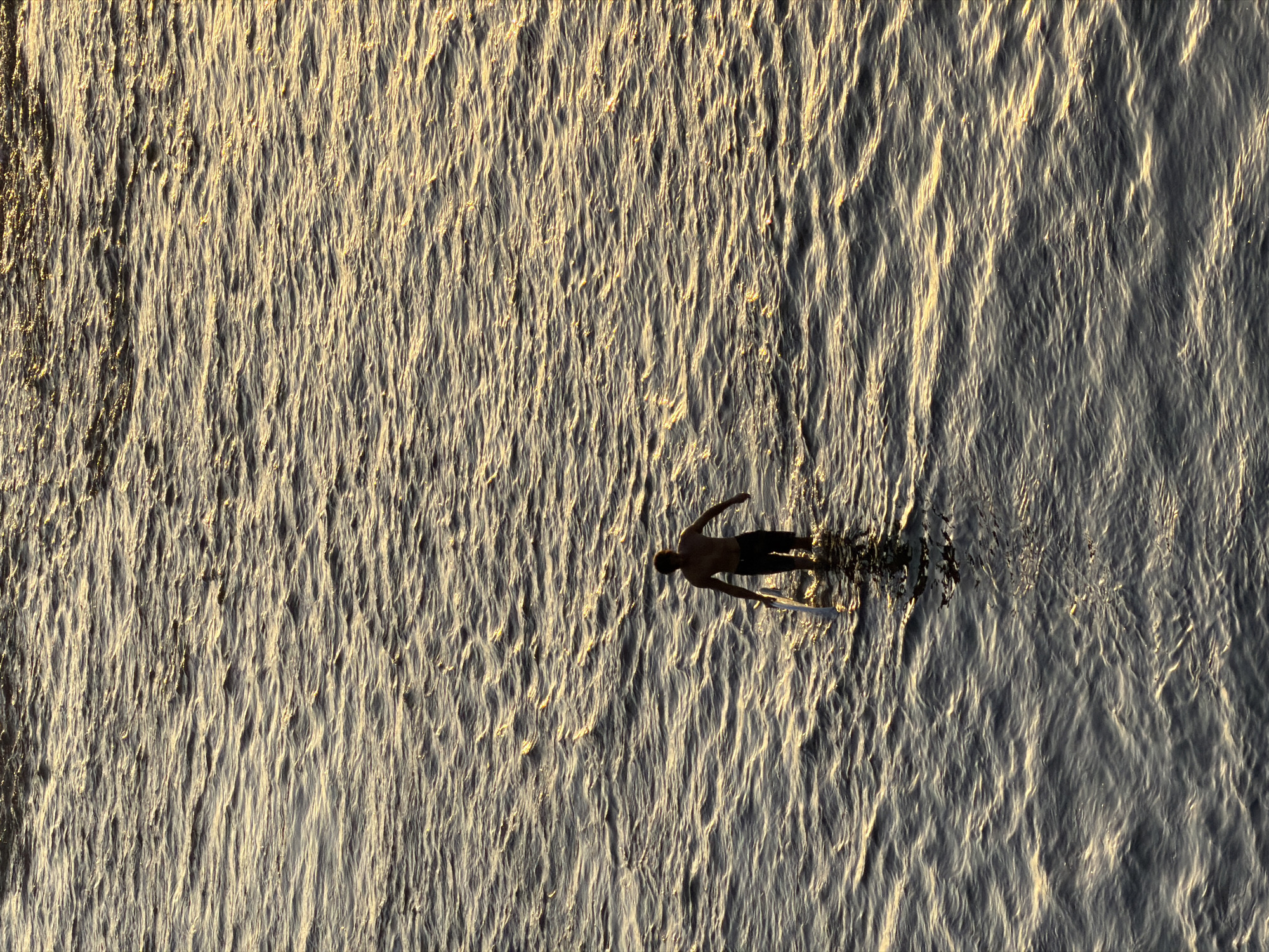 In the city of Kuta Selatan, Indonesia, a lone individual stands in the ocean, silhouetted against the golden light of a setting sun. The person is shirtless and wearing dark shorts, holding a surfboard by their side. The water, rippling with the gentle movement of the waves, reflects the warm hues of the sky, creating a shimmering, textured surface. The scene is captured from an elevated perspective, looking down on the solitary figure navigating the tranquil waters.