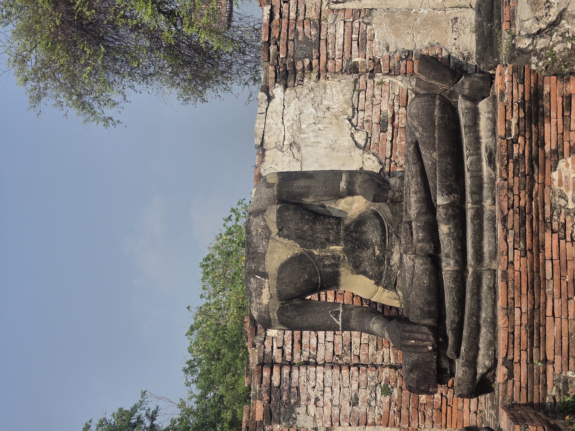 The image shows the torso of a Buddha statue at Wat Yai Chaimongkol, an ancient temple in Phra Nakhon Si Ayutthaya City Municipality, Thailand. The statue is made of stone and is missing its head. The torso is located adjacent to a red brick wall, with vegetation visible on either side. The sky is light blue.