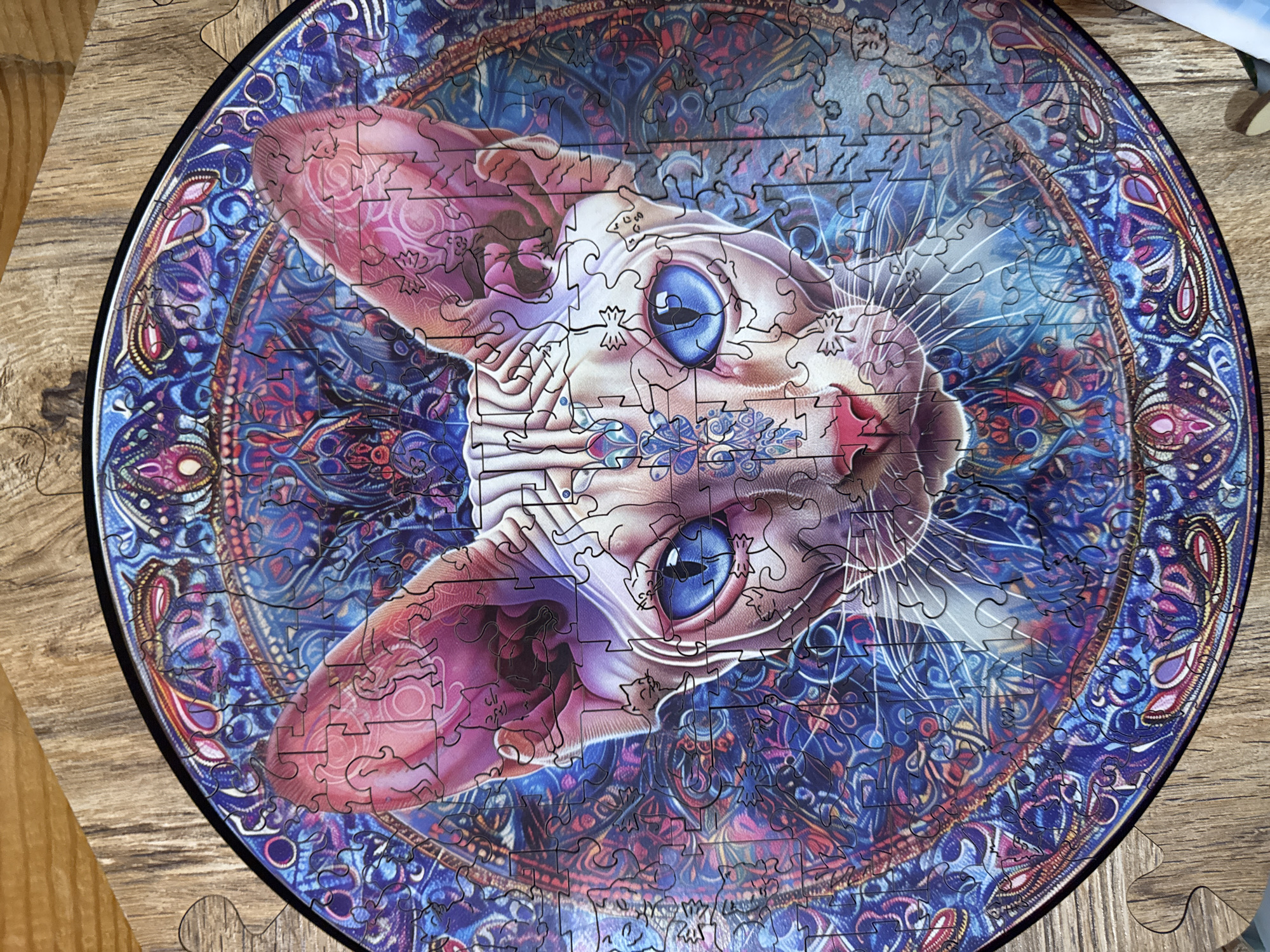 A circular jigsaw puzzle depicting a stylized Sphynx cat with vibrant blue eyes is laid on a wooden surface. The cat's face is the central focus, with intricate geometric patterns and swirling designs in shades of blue, purple, and pink surrounding it. Small, silhouetted cat figures are incorporated into the artwork on the puzzle. The puzzle itself is incomplete, with several pieces missing along the edges, revealing the wooden backing. The wooden surface appears to be a tabletop or floor with a visible wood grain texture. The image is a close-up, overhead shot of the puzzle. There is no indication of the city of Nikolaevka, Russia, in the image or its context.